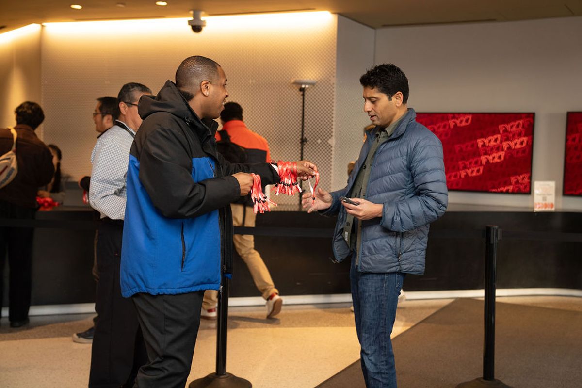 Two men exchange a gift bag in a lobby area with red screens and other attendees in the background.
