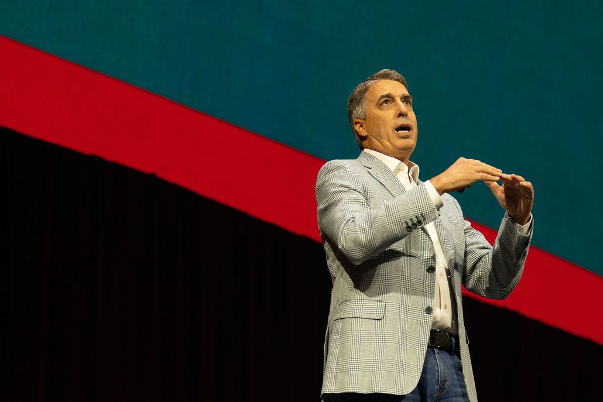 Speaker in gray suit gesturing while presenting on stage with red and teal geometric backdrop