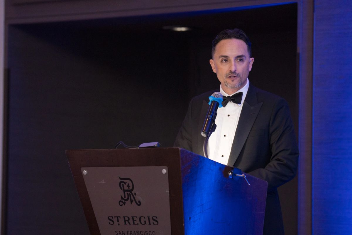 Speaker in black tuxedo presenting at podium with St. Regis logo at formal evening event