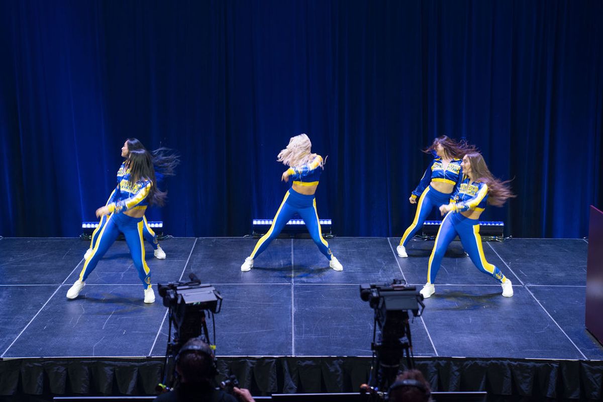 Three cheerleaders in blue and yellow uniforms performing synchronized dance routine on stage with blue.