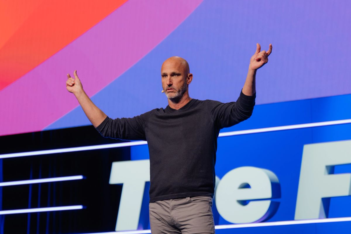 Bald speaker in black shirt gestures with both arms raised on stage with colorful geometric backdrop