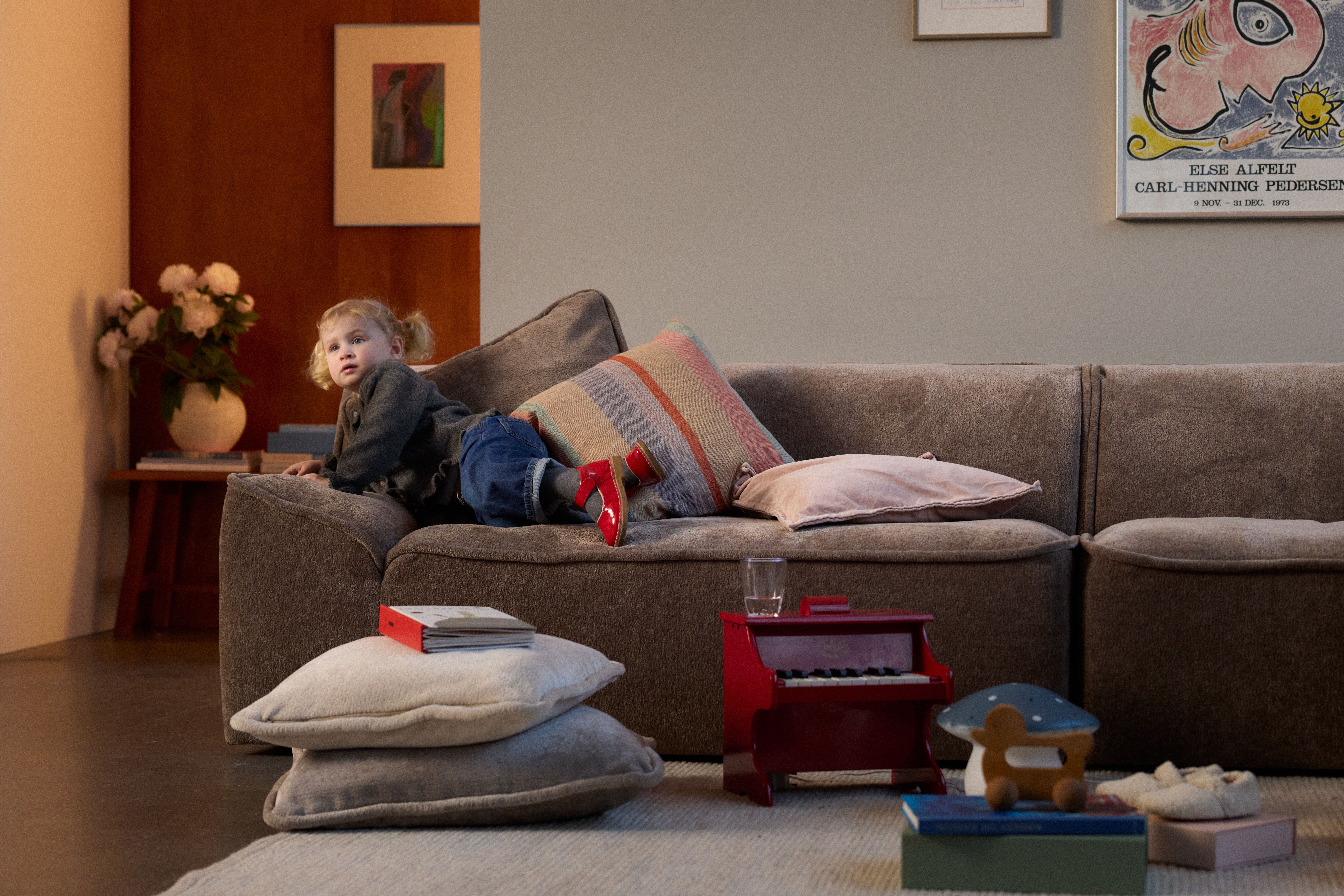 A young child lies on a brown sofa, looking at the viewer, with a red toy piano and other toys on the floor.