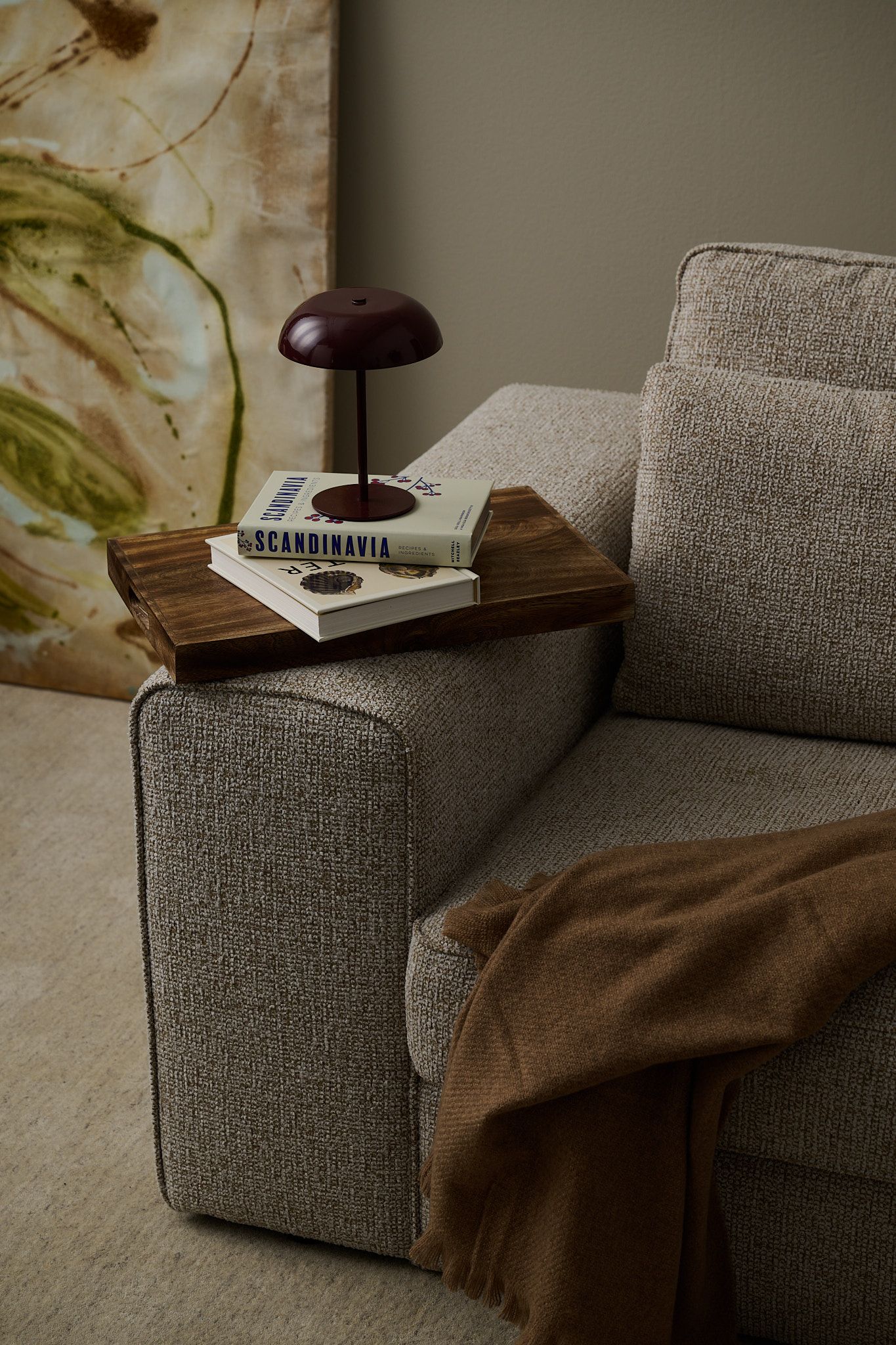 A textured light grey sofa with a wooden armrest tray holding two books and a red mushroom lamp, and a brown throw blanket.