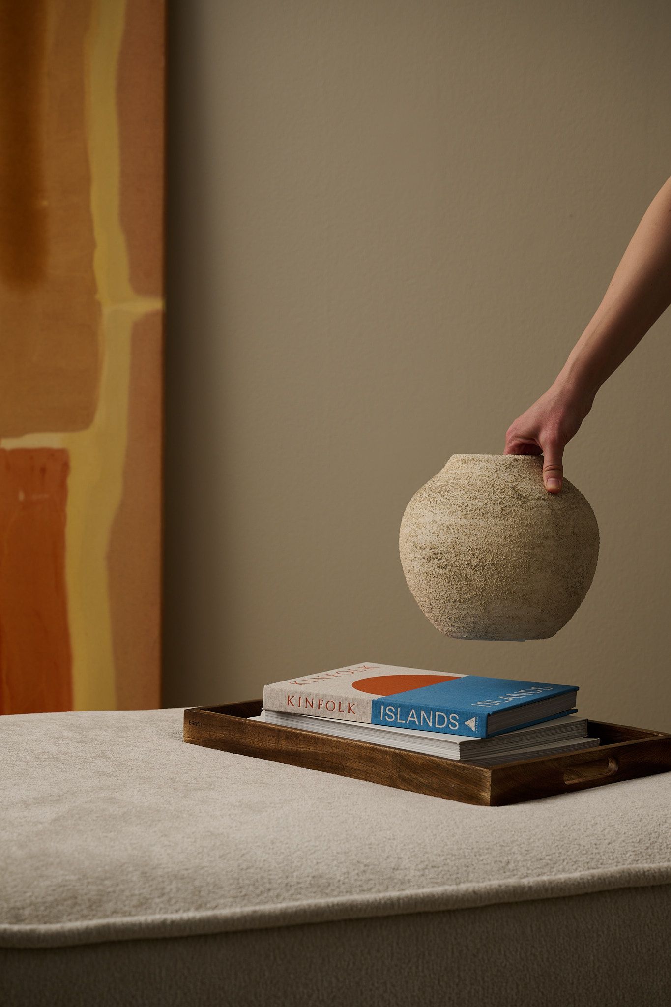 A hand holds a textured vase above books on a wooden tray, resting on a light upholstered surface, with abstract art in the background.