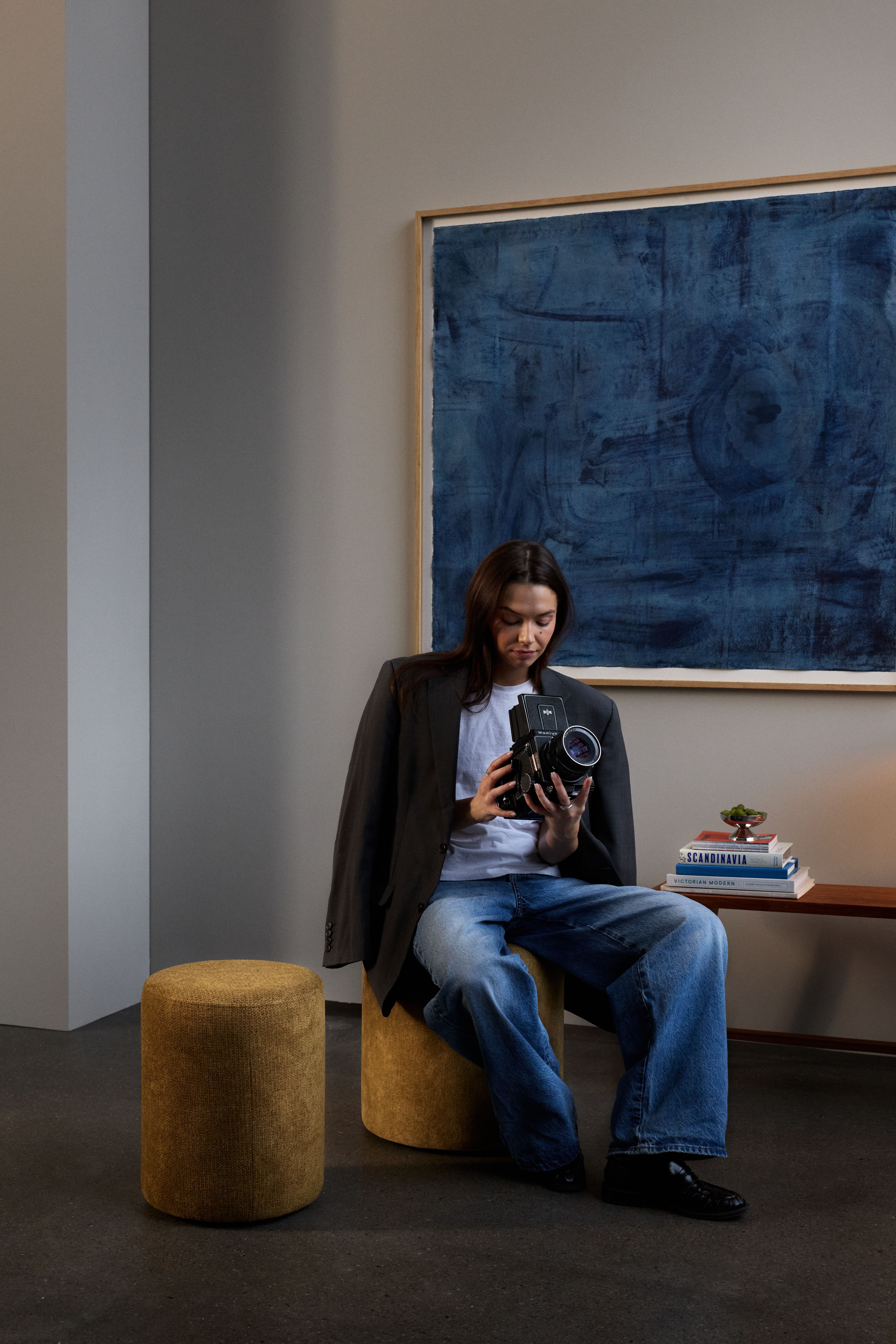 A person wearing a blazer, white t-shirt, and jeans sits on a yellow stool, looking at a camera. Behind them, a large abstract blue painting hangs on a gray wall.