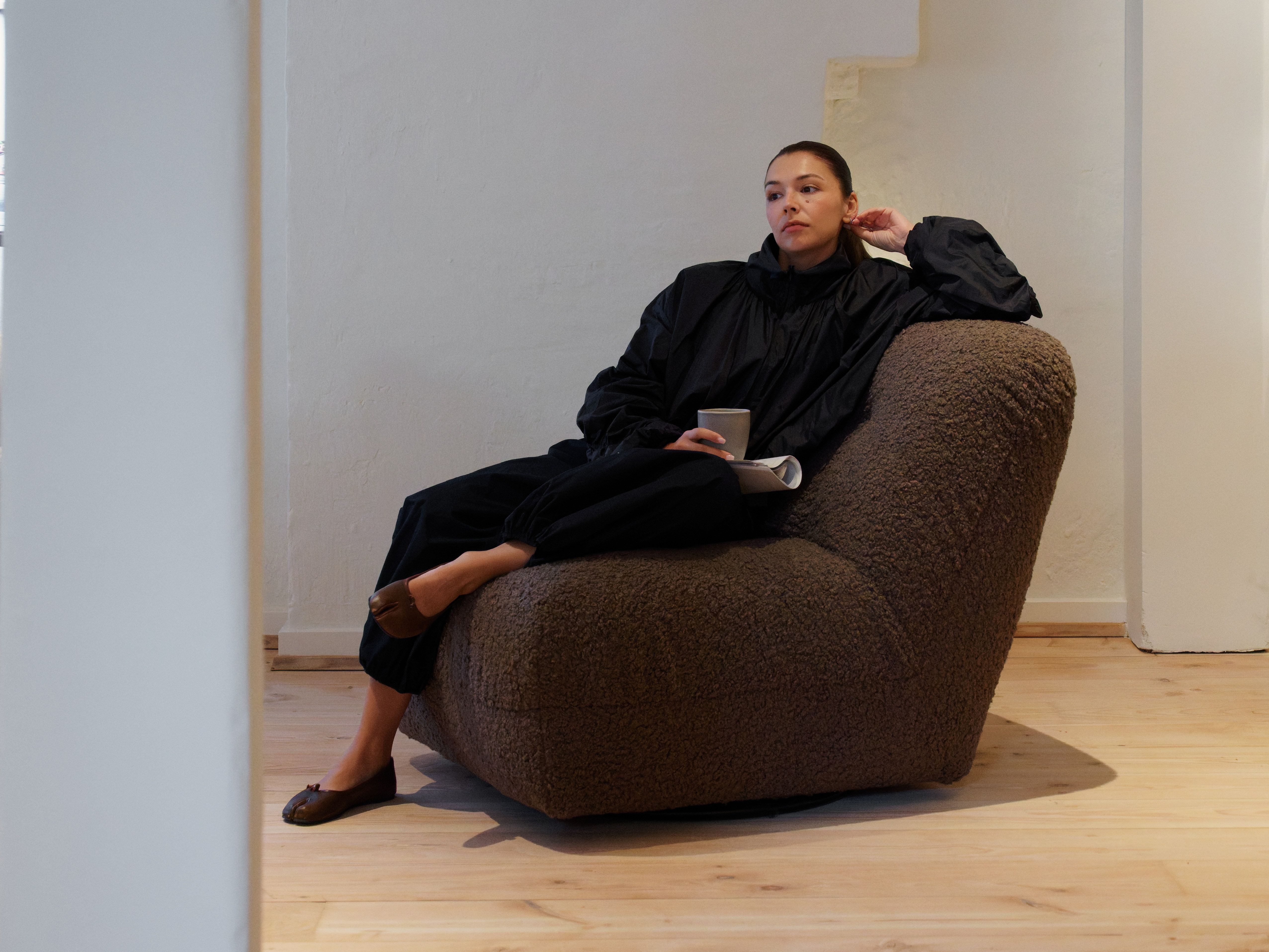 A woman in a black jacket sits thoughtfully in a brown textured chair, holding a cup.
