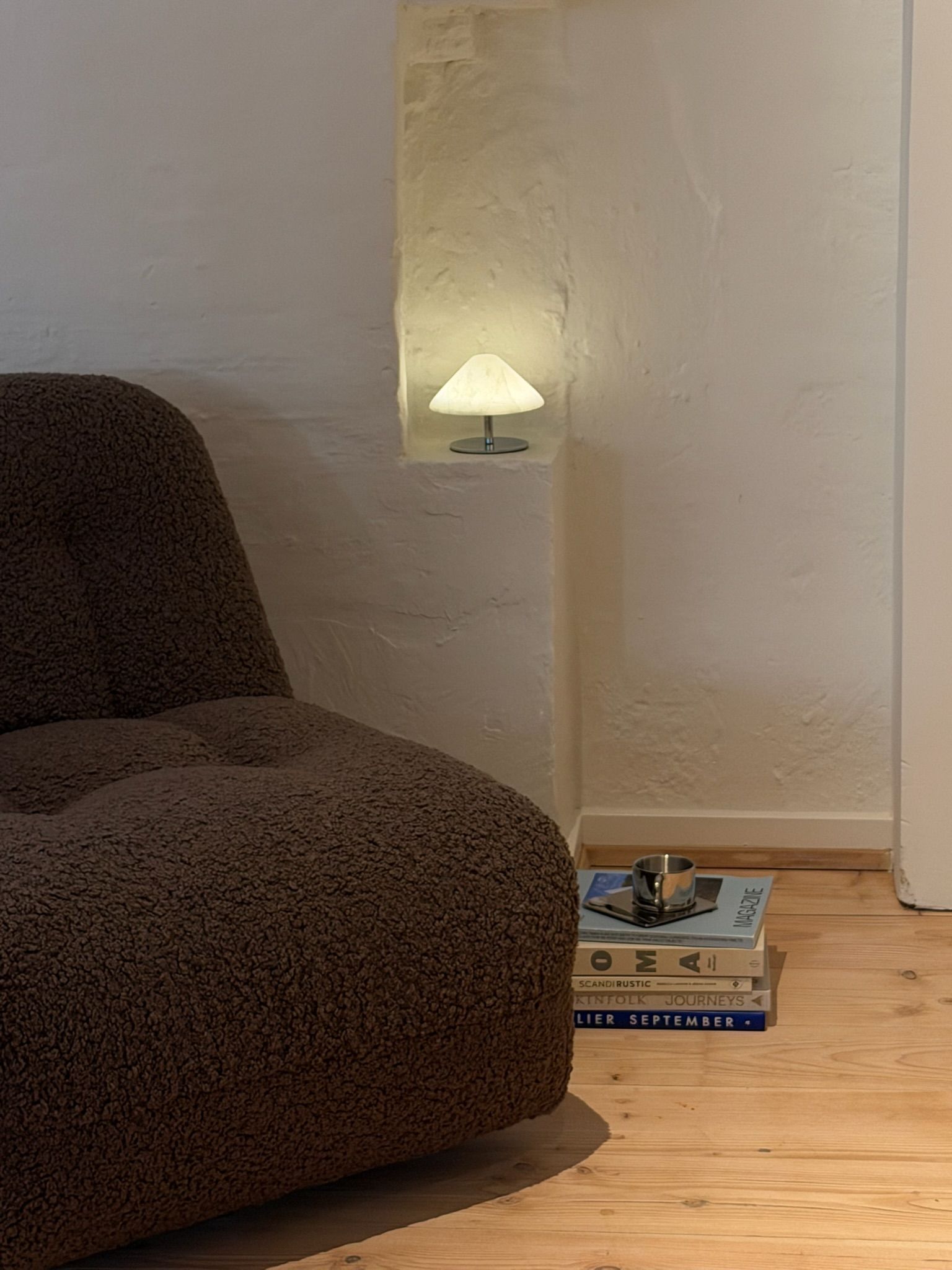 Brown sherpa armchair next to a white wall niche with a glowing mushroom lamp and a stack of books on a wooden floor.