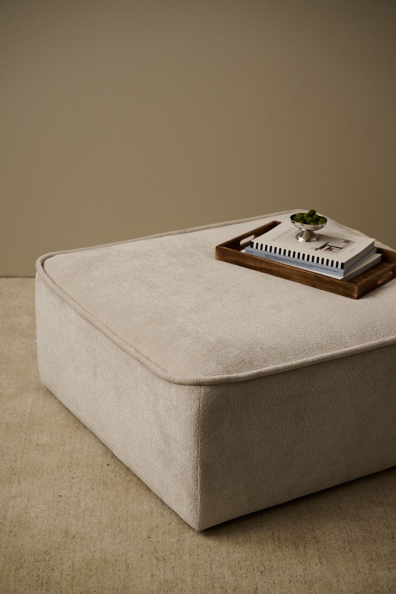 A light beige rectangular ottoman with a wooden tray holding books and a silver bowl of green spheres.
