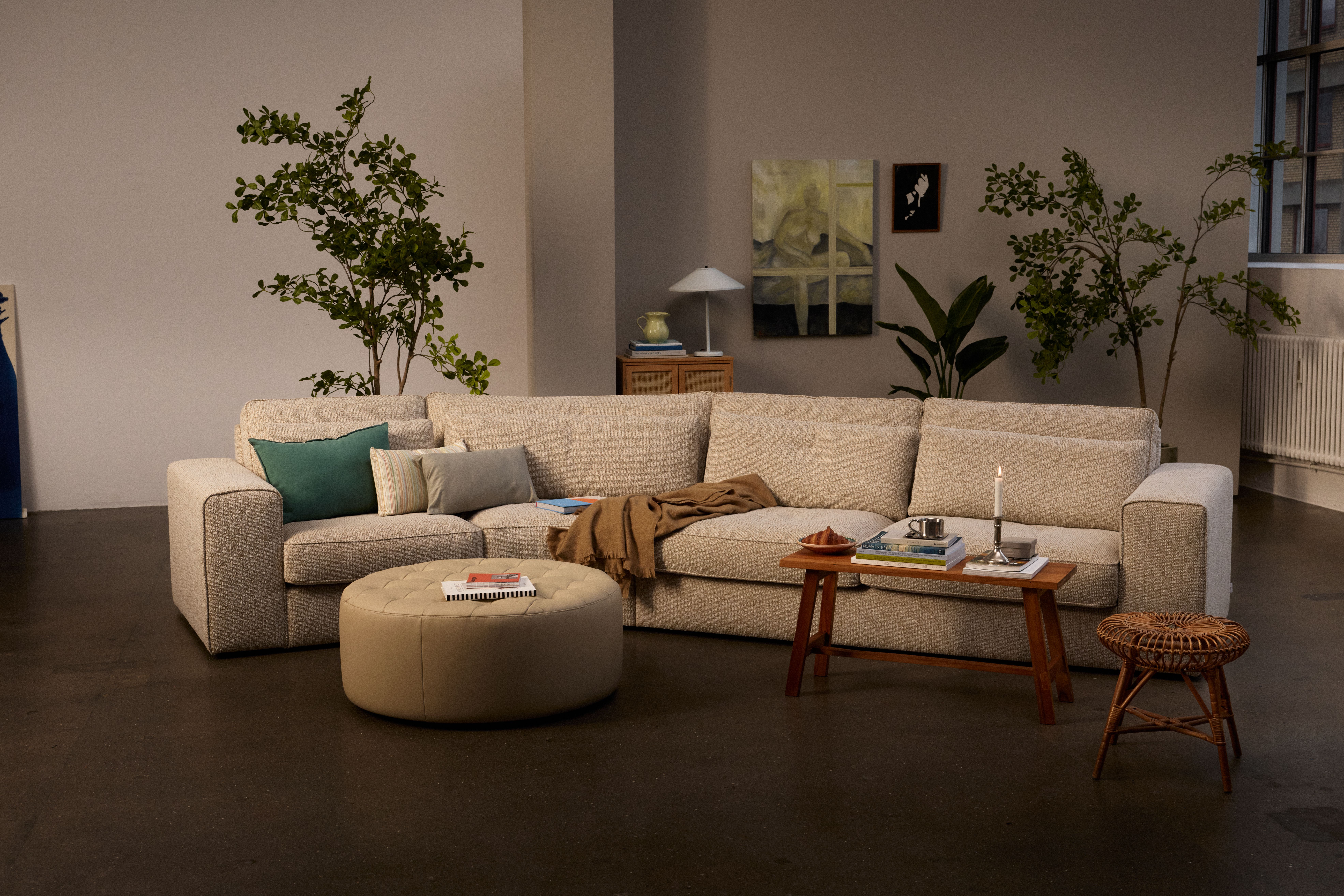 A modern living room with a large cream sectional sofa, a round ottoman, and tall green plants.