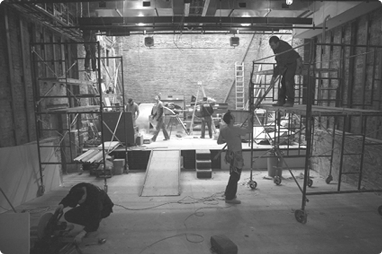 A wide shot of the theater stage being set up. Workers move around while scaffolds are set up on either side. 