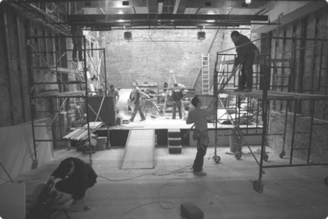 A wide shot of the theater stage being set up. Workers move around while scaffolds are set up on either side.