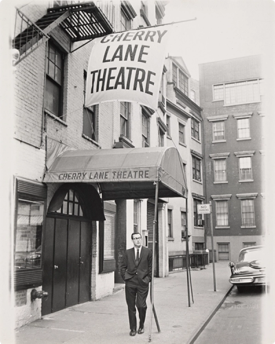 A black and white photo of the exterior of the Cherry Lane theater. The awning and banner both blow in the breeze, displaying the theater's name. A man in a suit leans against the railing. 