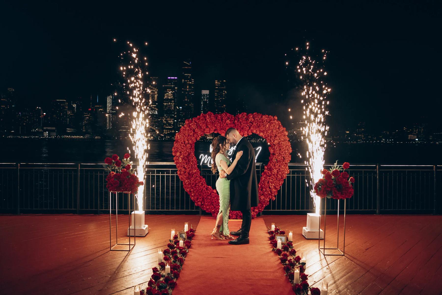 Couple embracing at night in front of a floral installation by the waterfront.