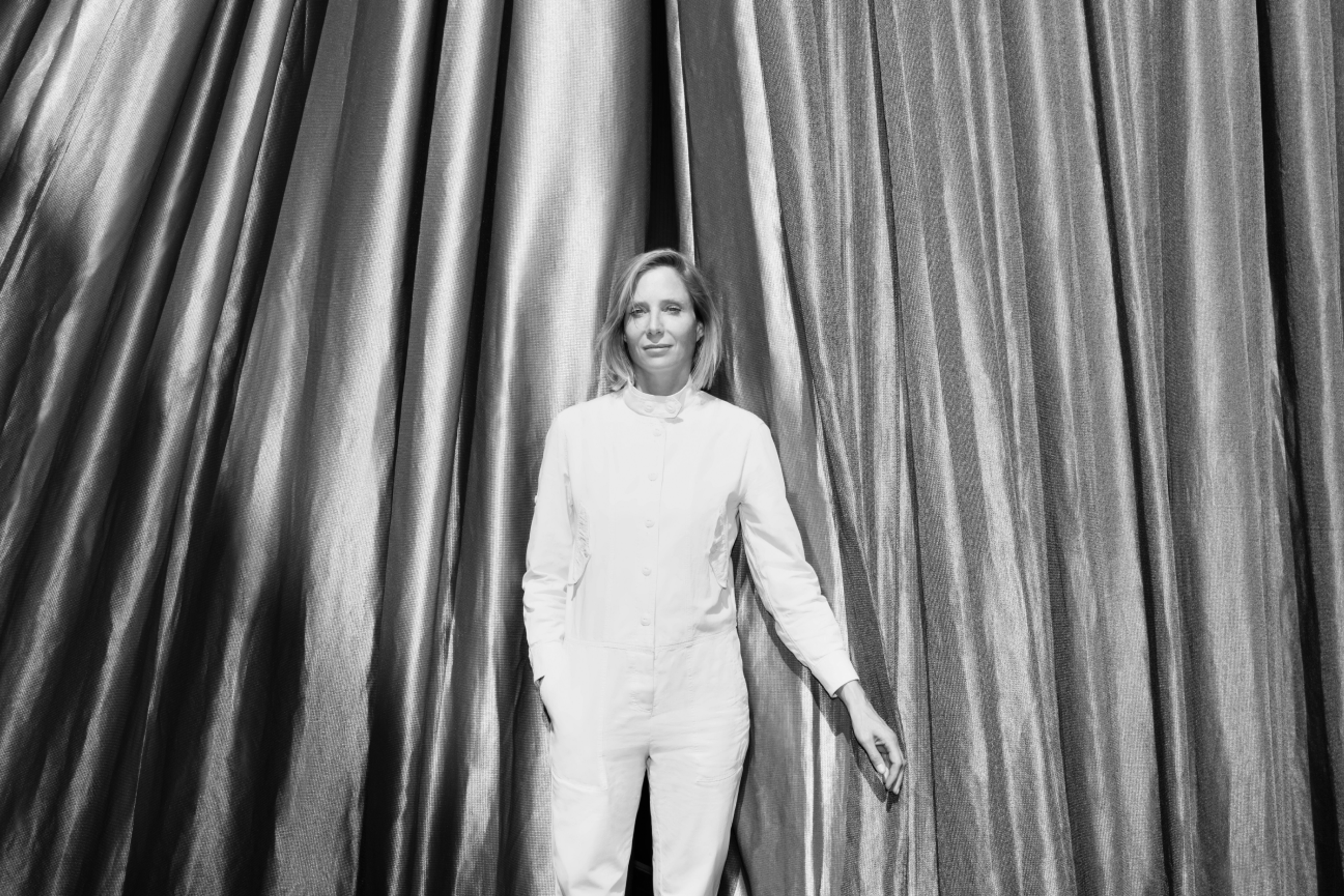 Black-and-white portrait of Dr. Bettina Kames, director of the Berlin-based art foundation LAS, standing in front of curtains.