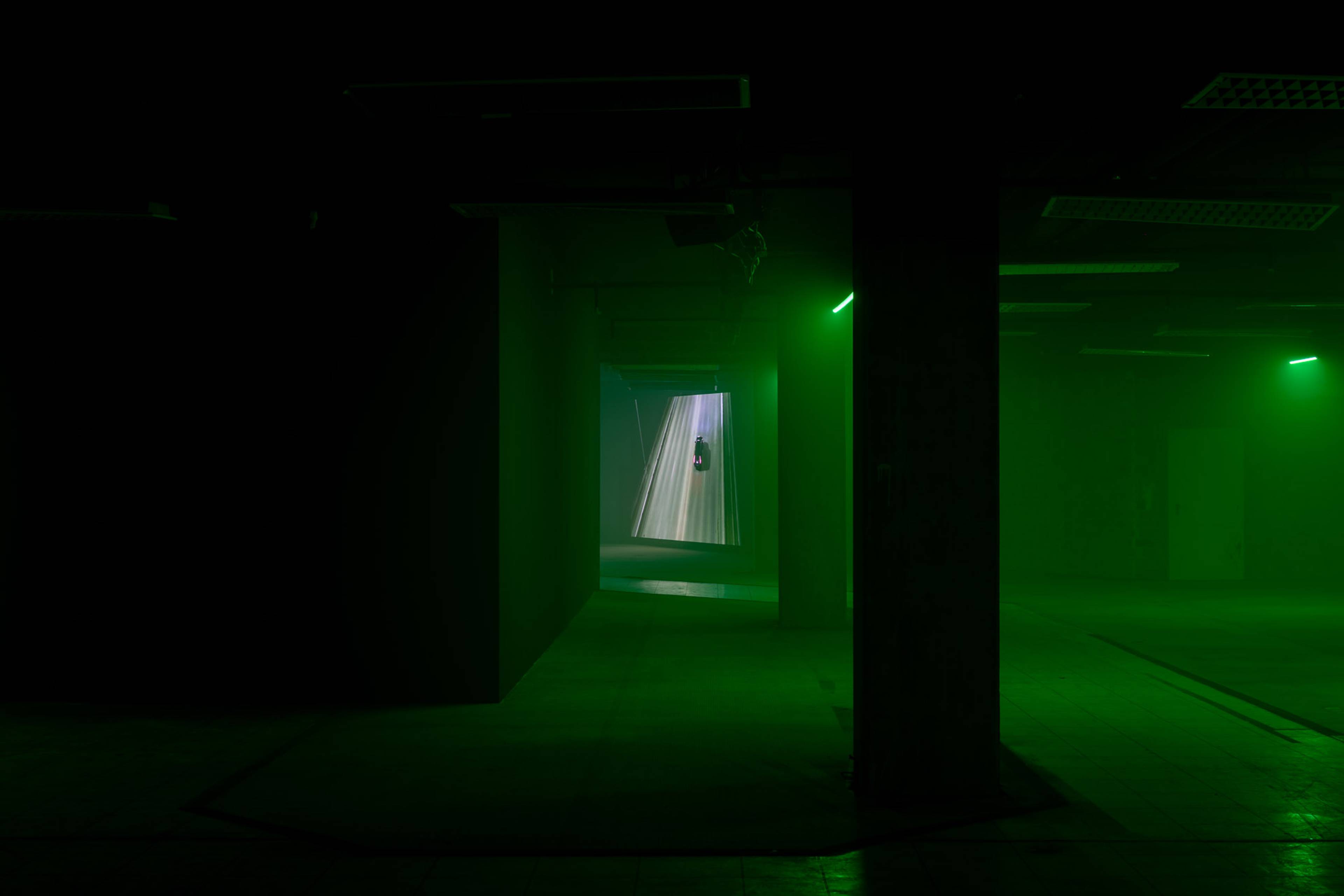 Dunkler Raum mit grüner Beleuchtung, Blick durch einen Durchgang auf eine Projektion, die eine schwebende Figur in vertikal verlaufenden Lichtstrahlen zeigt.