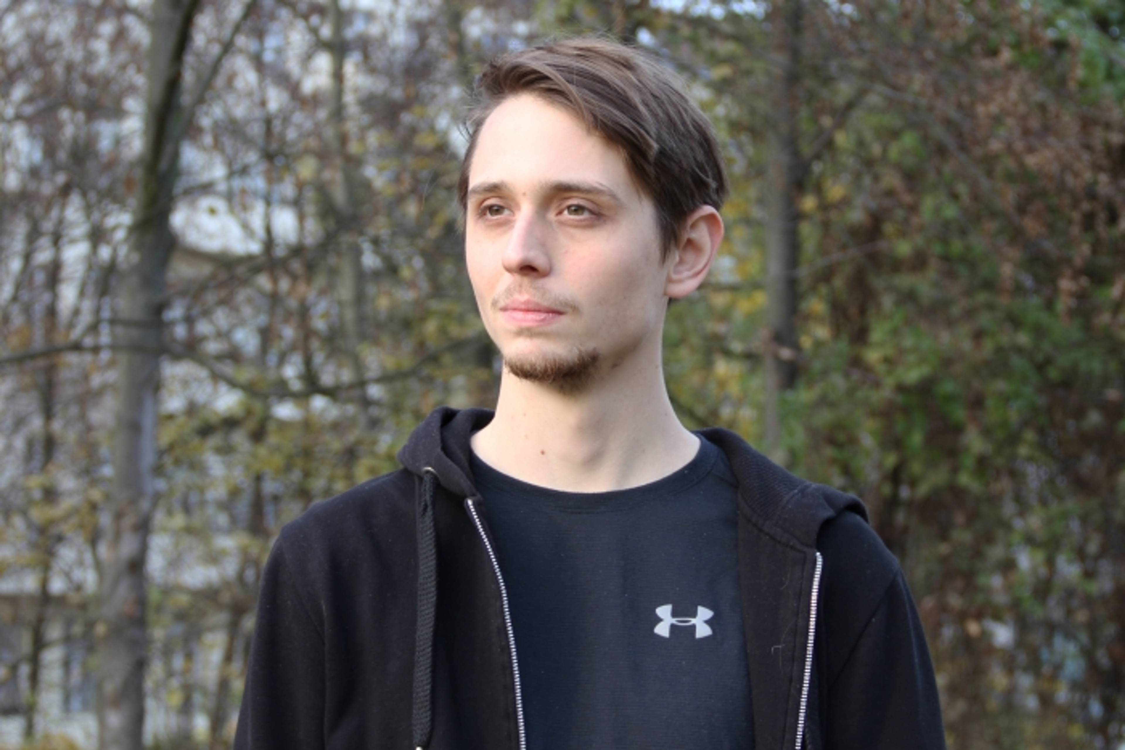 A person with short hair stands outdoors, wearing a black hoodie over a black Under Armour shirt. The background features trees with fall foliage.