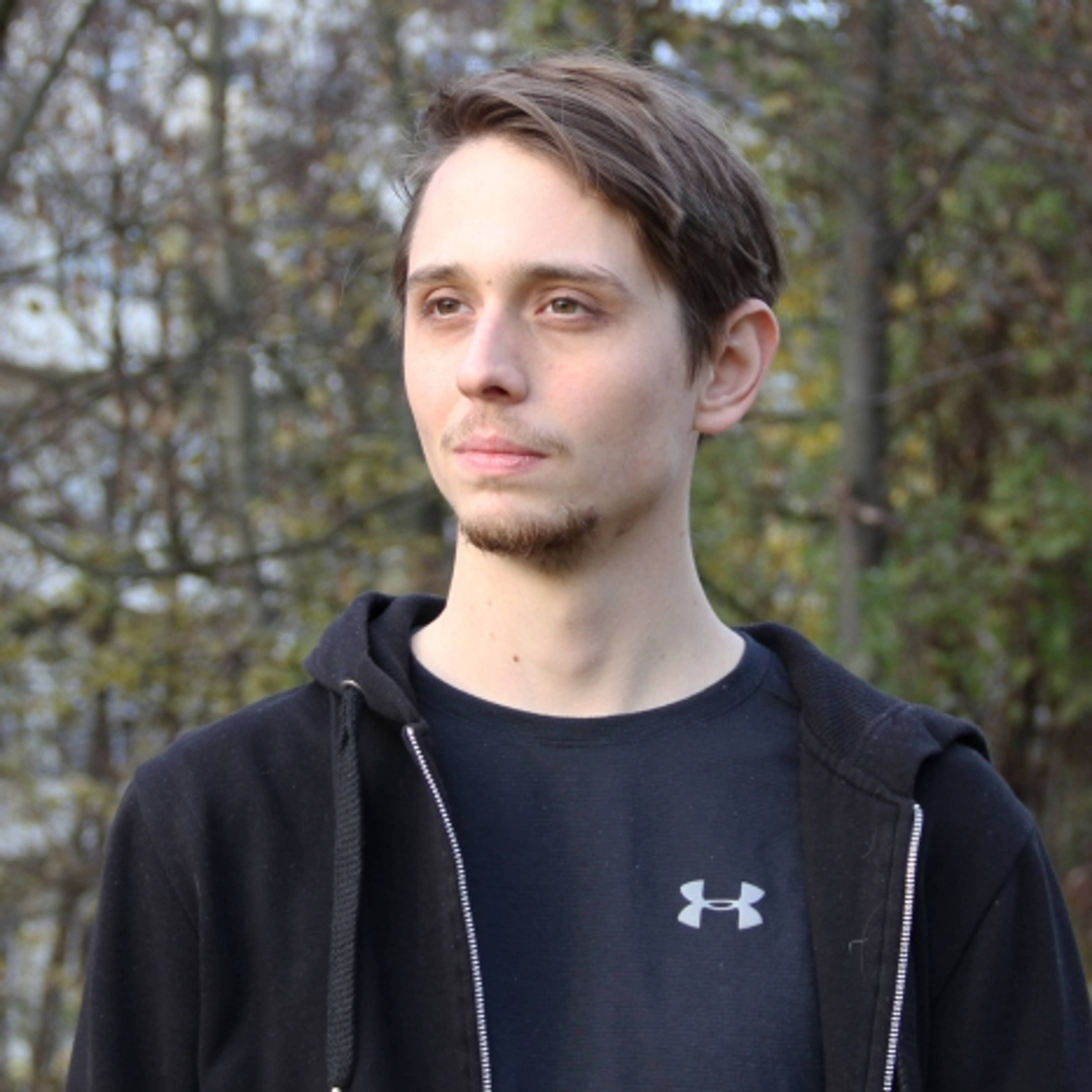 A person with short hair stands outdoors, wearing a black hoodie over a black Under Armour shirt. The background features trees with fall foliage.
