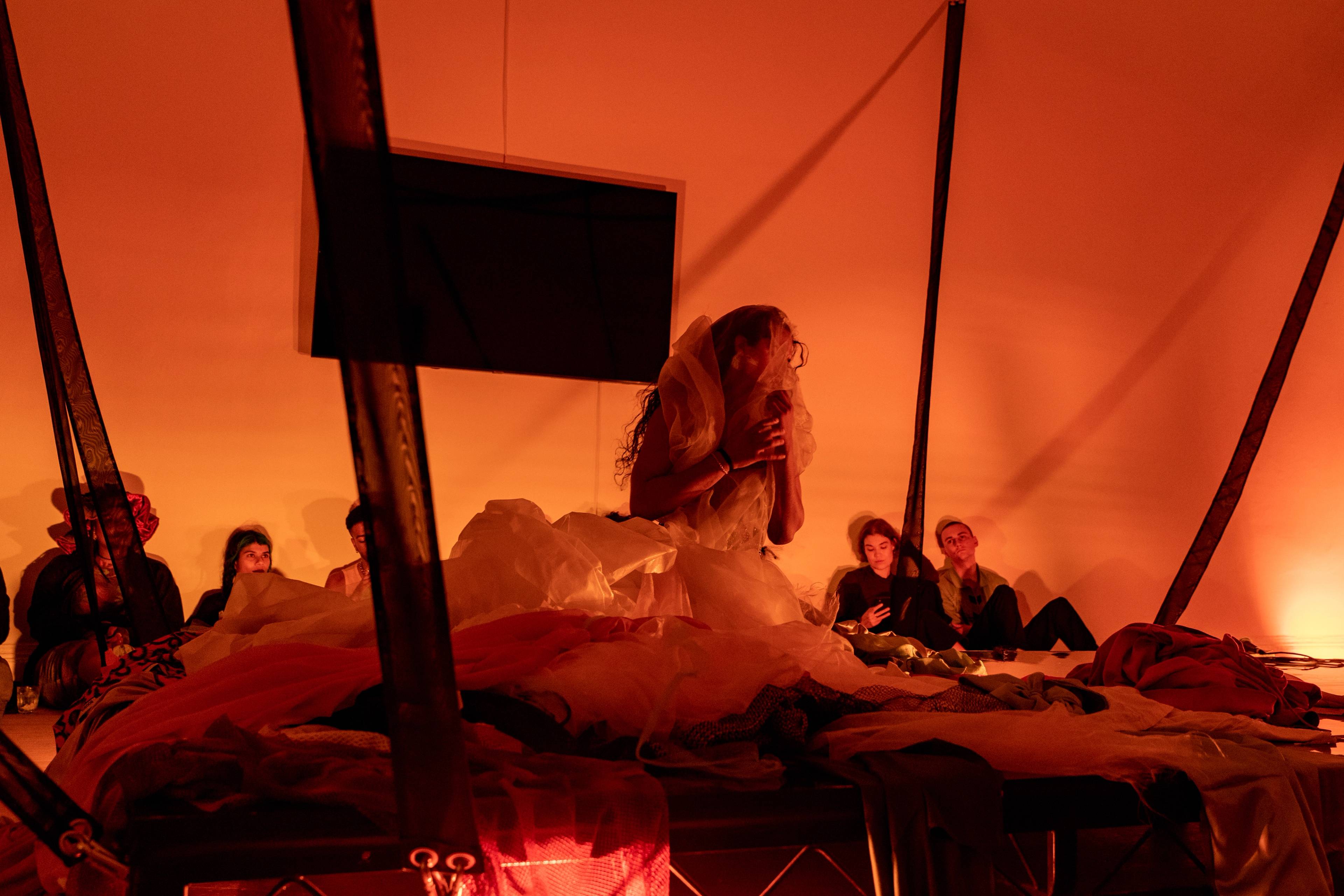 Eine Performer:in in einem weißen, mehrlagigen Outfit sitzt auf einer mit Stoffen bedeckten Plattform und hält einen Schleier vors Gesicht, während Zuschauer:innen im Hintergrund im warmen, orangefarbenen Licht sitzen.