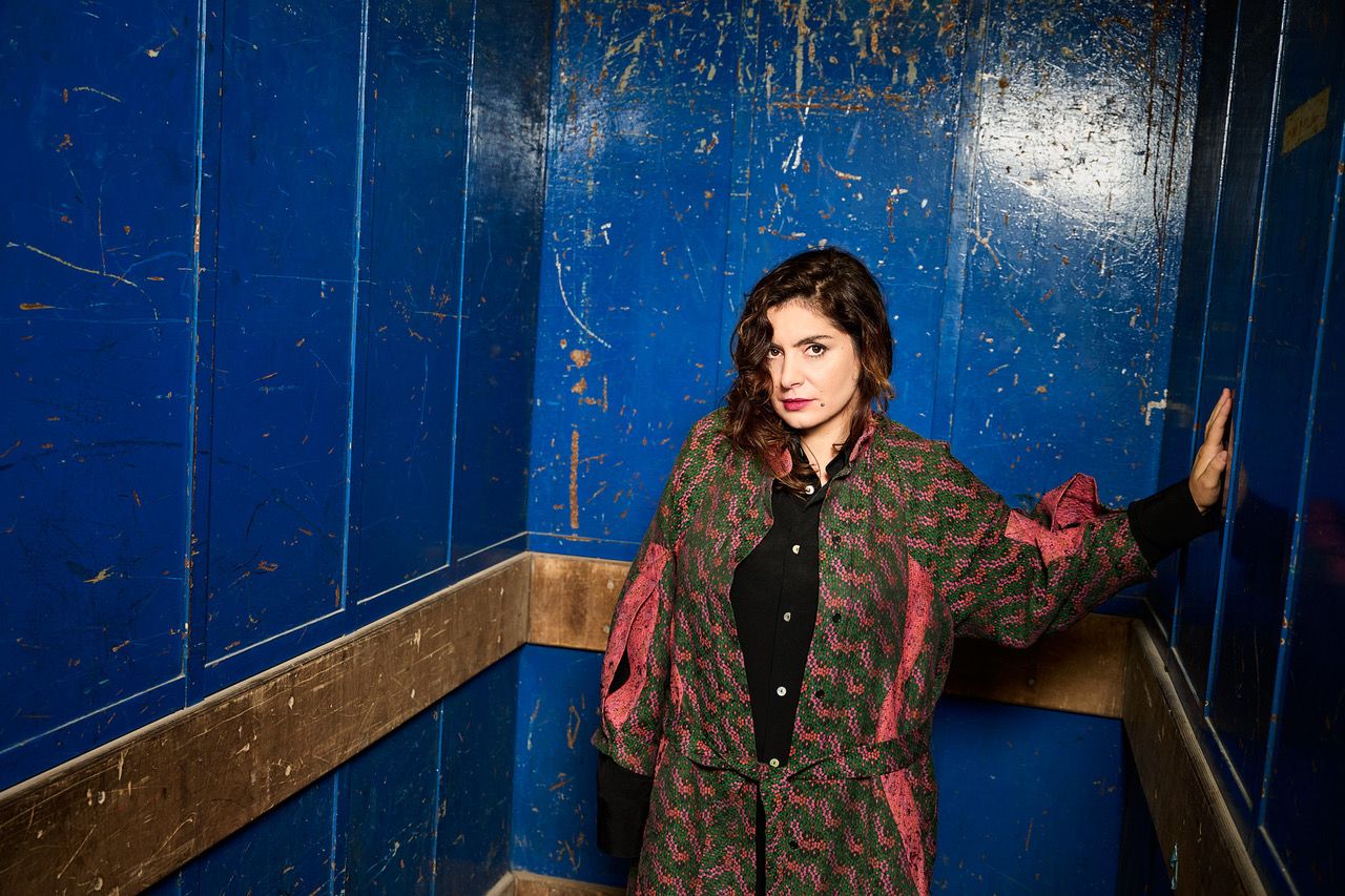 A person with shoulder-length dark hair stands in a blue room, wearing a patterned red and green coat over a black shirt, looking toward the camera.