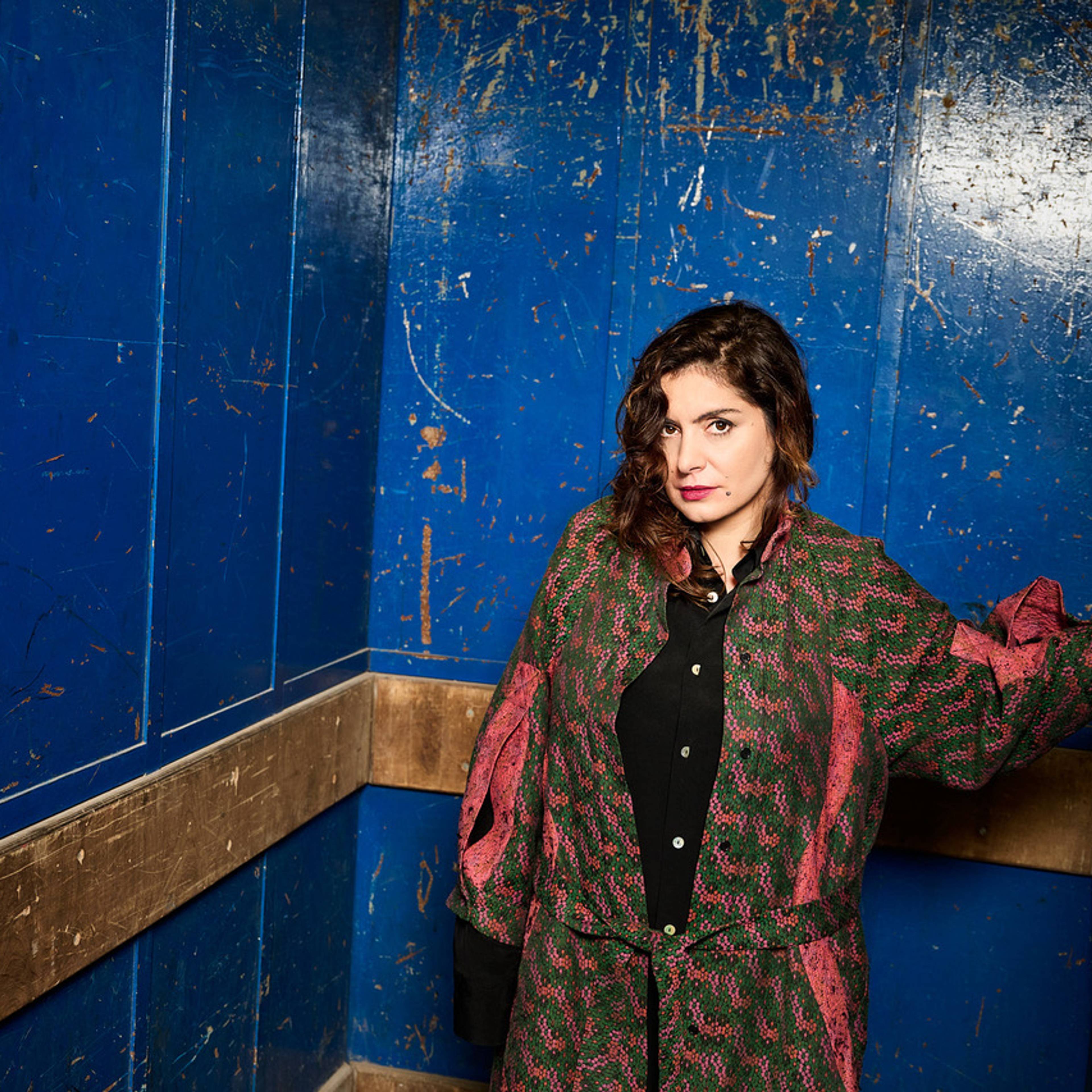 A person with shoulder-length dark hair stands in a blue room, wearing a patterned red and green coat over a black shirt, looking toward the camera.