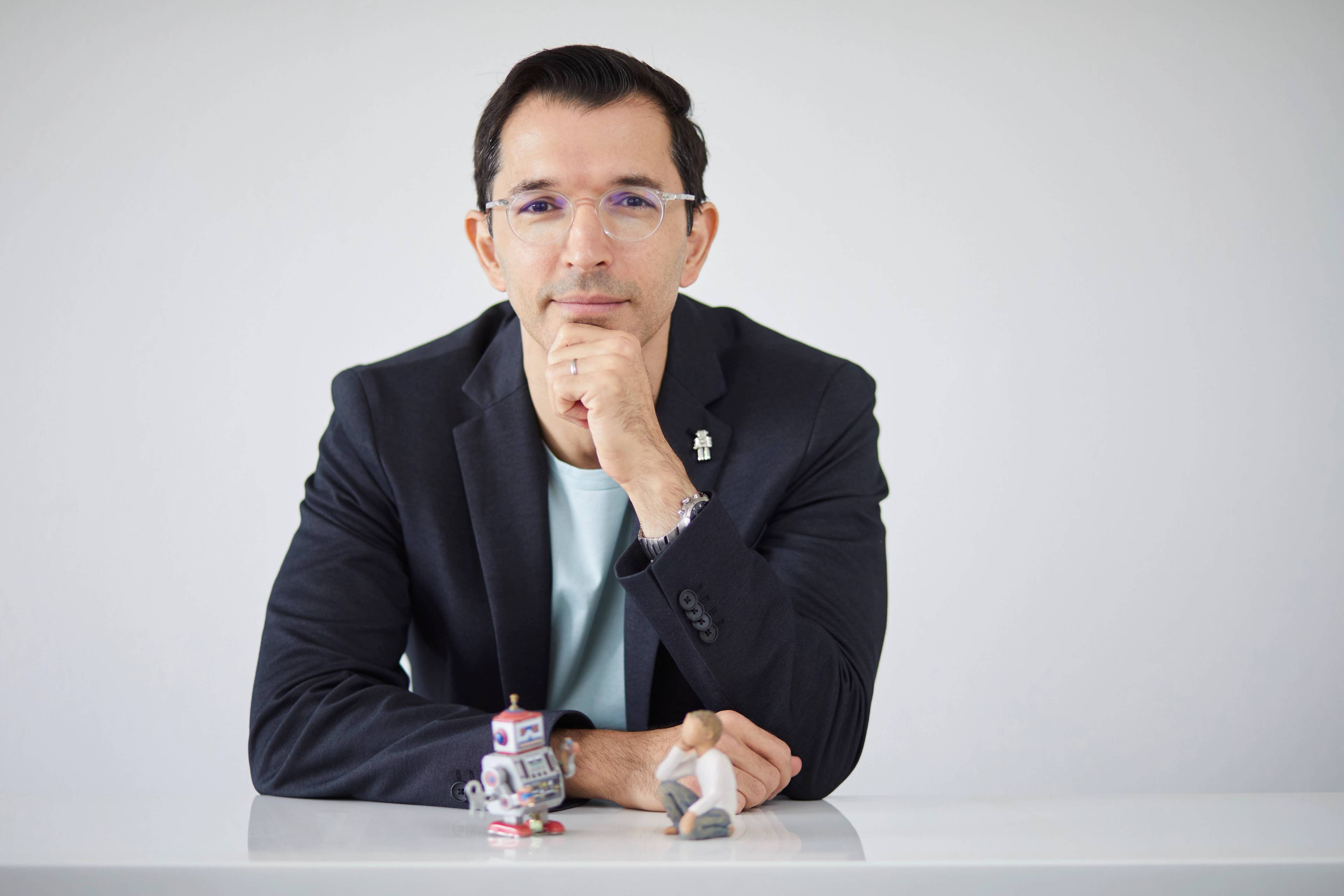 Person wearing glasses and a blazer, resting their chin on their hand, seated at a white table with small figurines of a robot and a miniature seated figure in front of them, against a plain background.