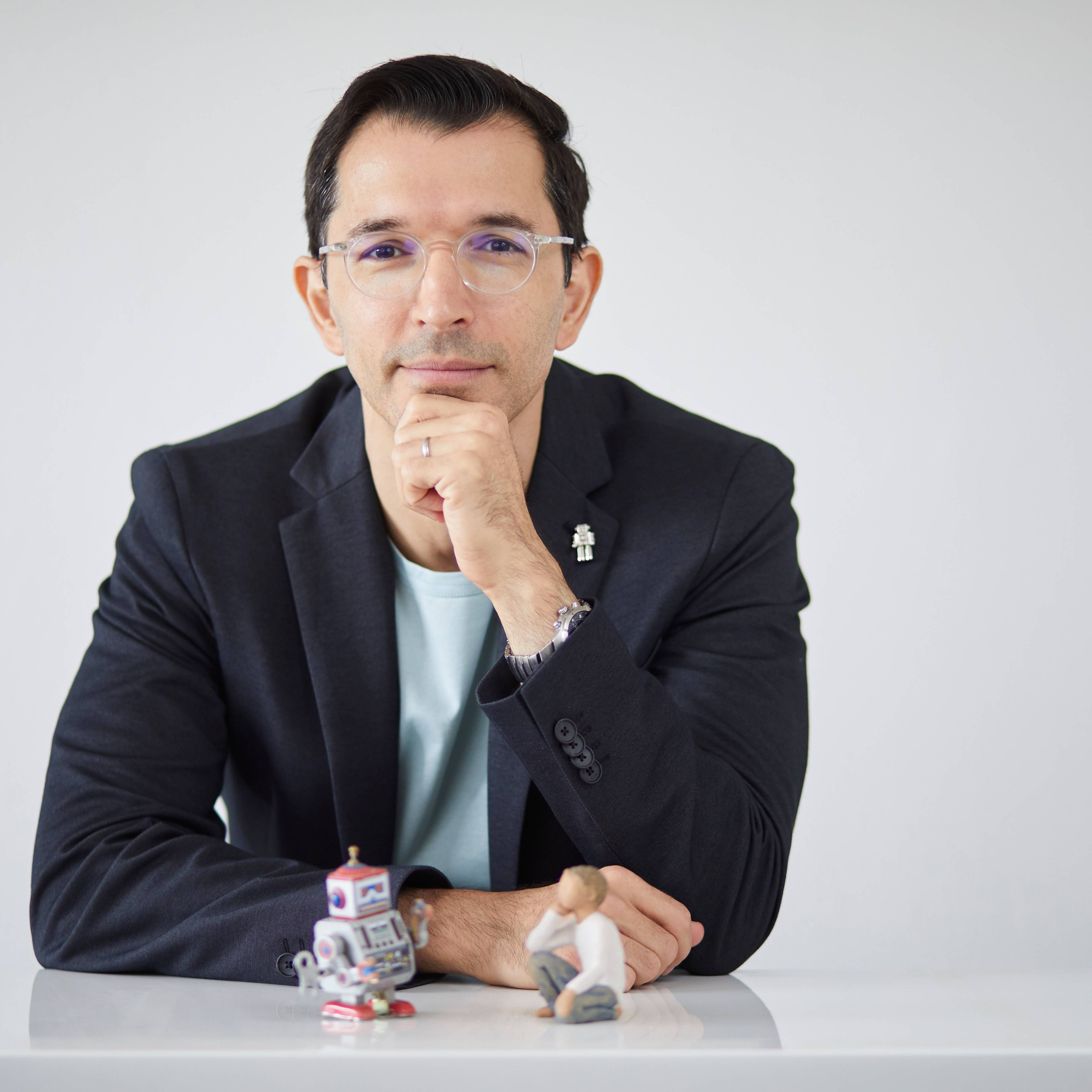 Person mit Brille und Blazer, stützt das Kinn auf die Hand, sitzt an einem weißen Tisch mit kleinen Figuren eines Roboters und einer sitzenden Miniaturfigur vor sich, vor einem schlichten Hintergrund.