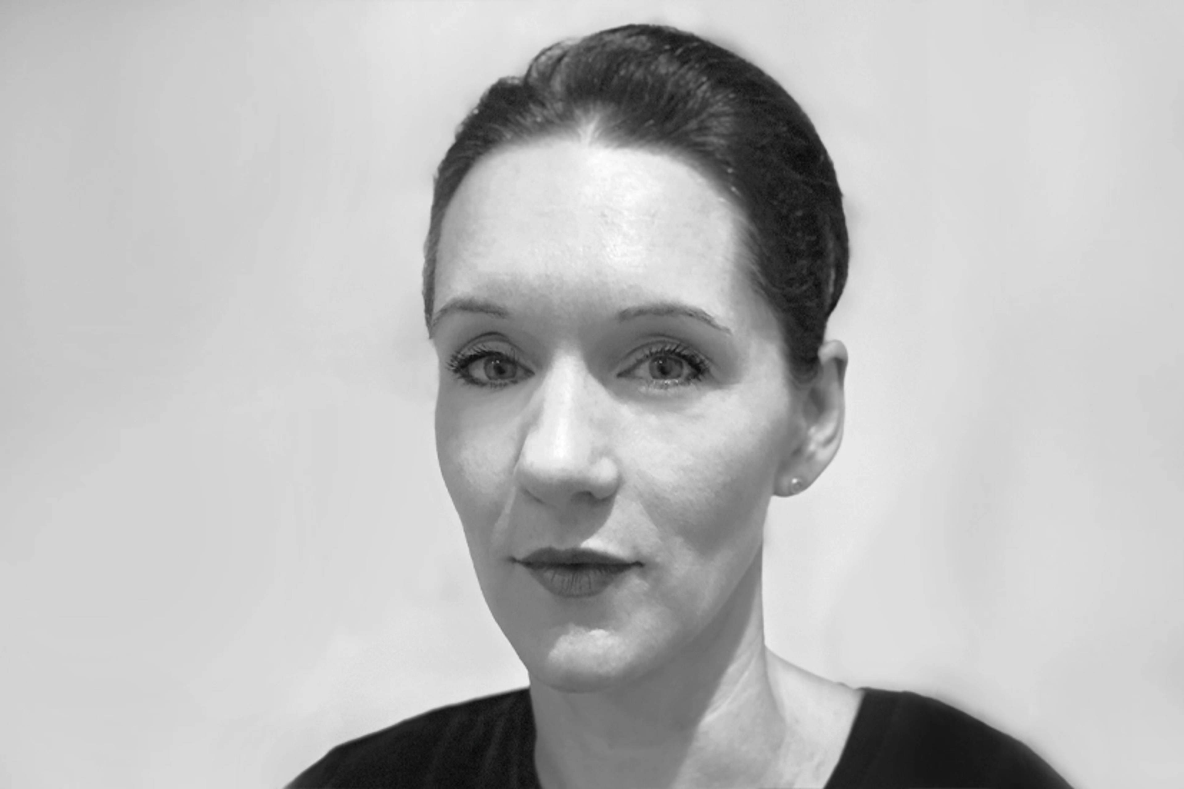 Black and white portrait of a woman with her hair pulled back, wearing a dark top against a plain light background. She is looking directly at the camera with a neutral expression.