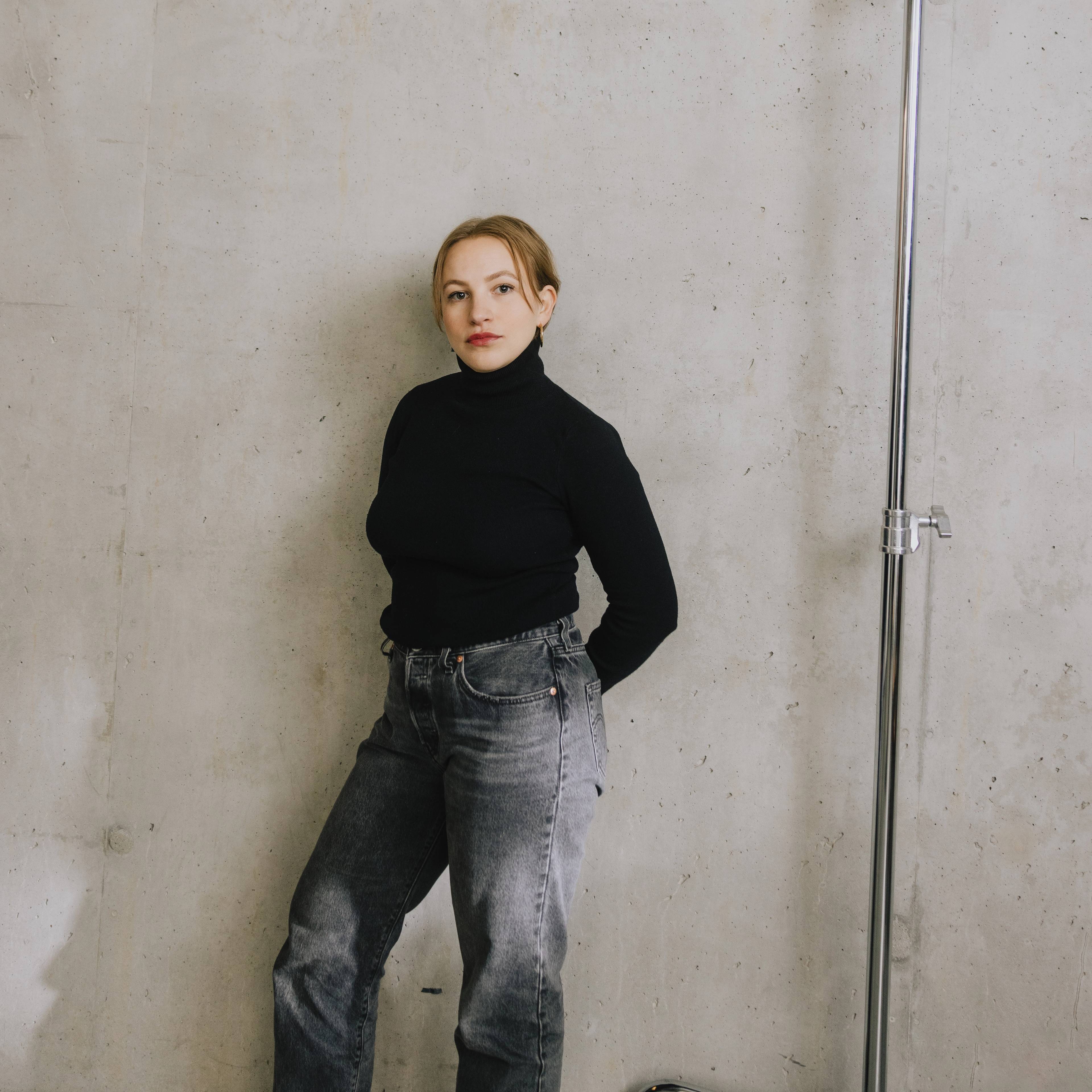 Portrait of Agnessa Schmudke, Assistant Curator and Lead of Live and Outreach Programmes at LAS, standing against a concrete wall in a black turtleneck and grey denim trousers in a minimalist studio setting.