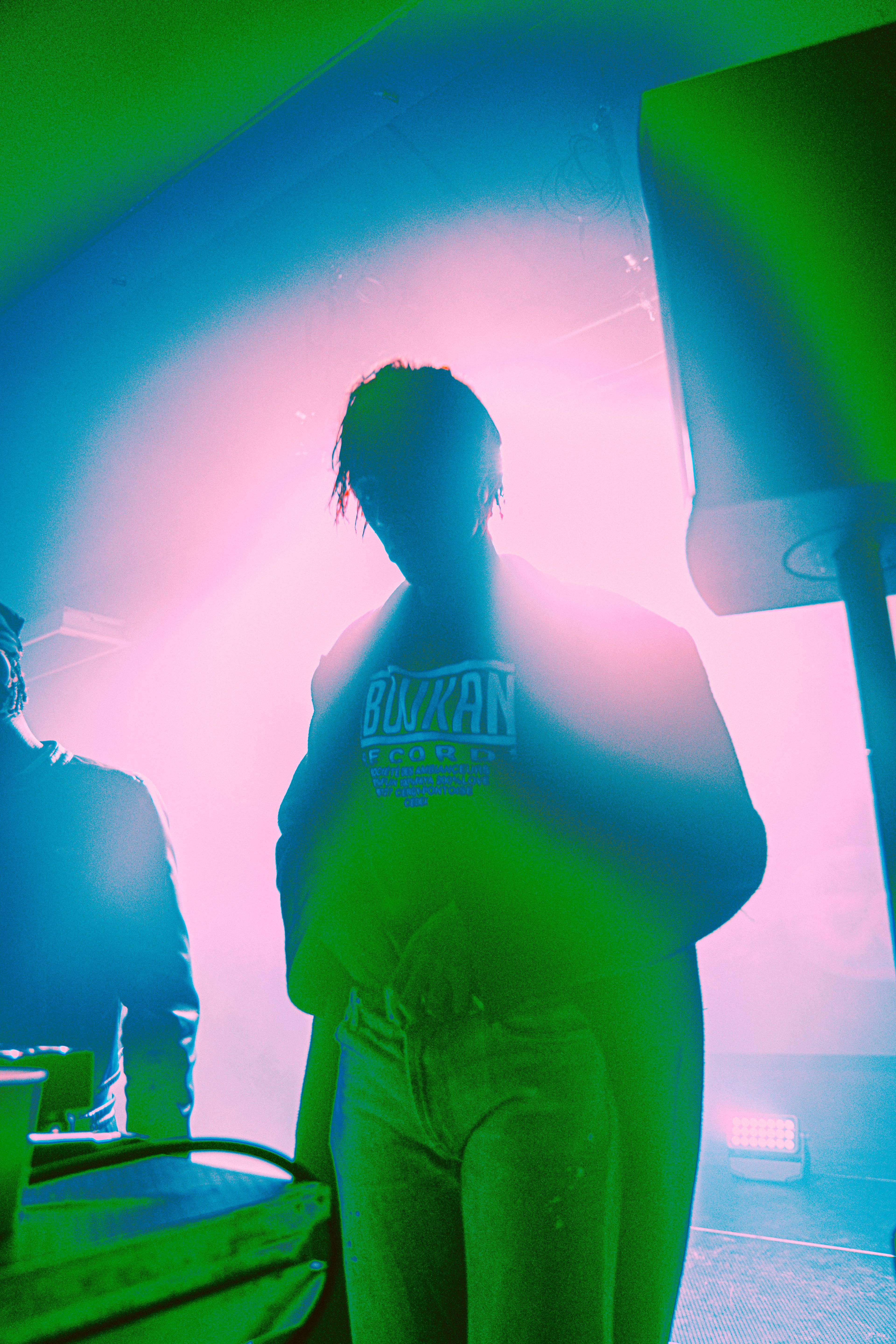 A performer in a layered white outfit sits on a platform surrounded by fabric, holding a veil to their face, while people watch from the background in dim orange lighting.