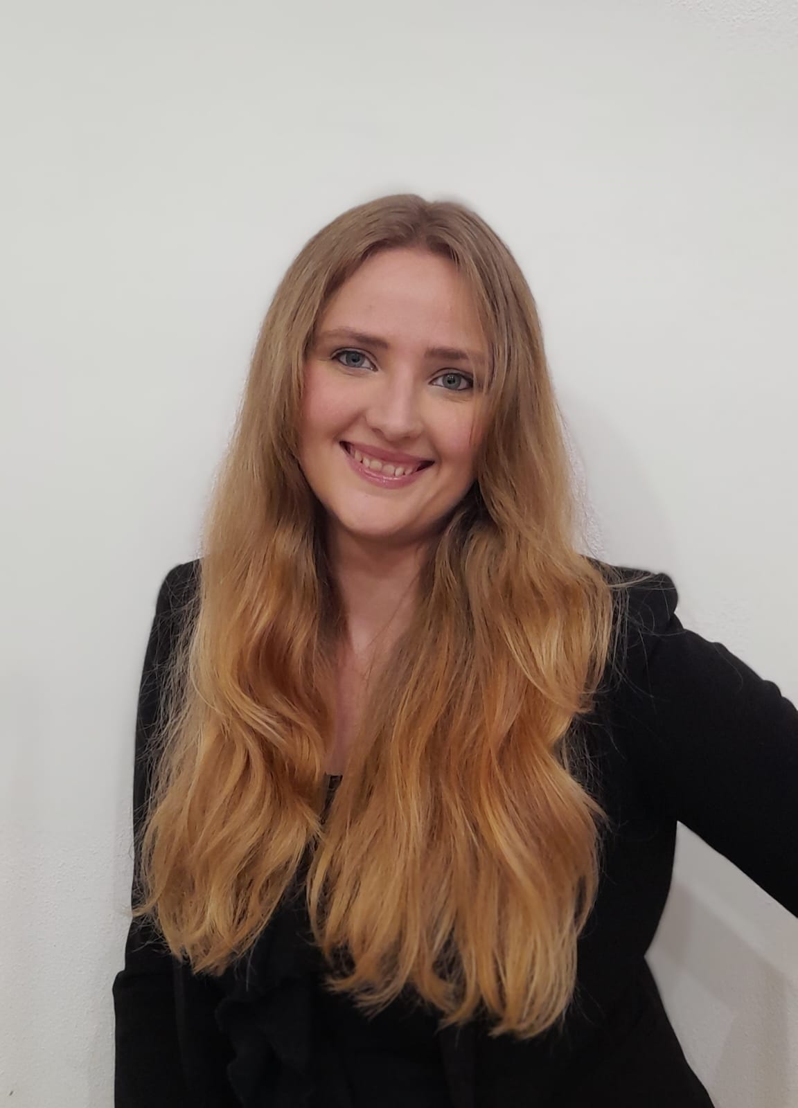 A person with long, wavy blonde hair is smiling warmly at the camera. They are wearing a black top and standing against a plain white background.