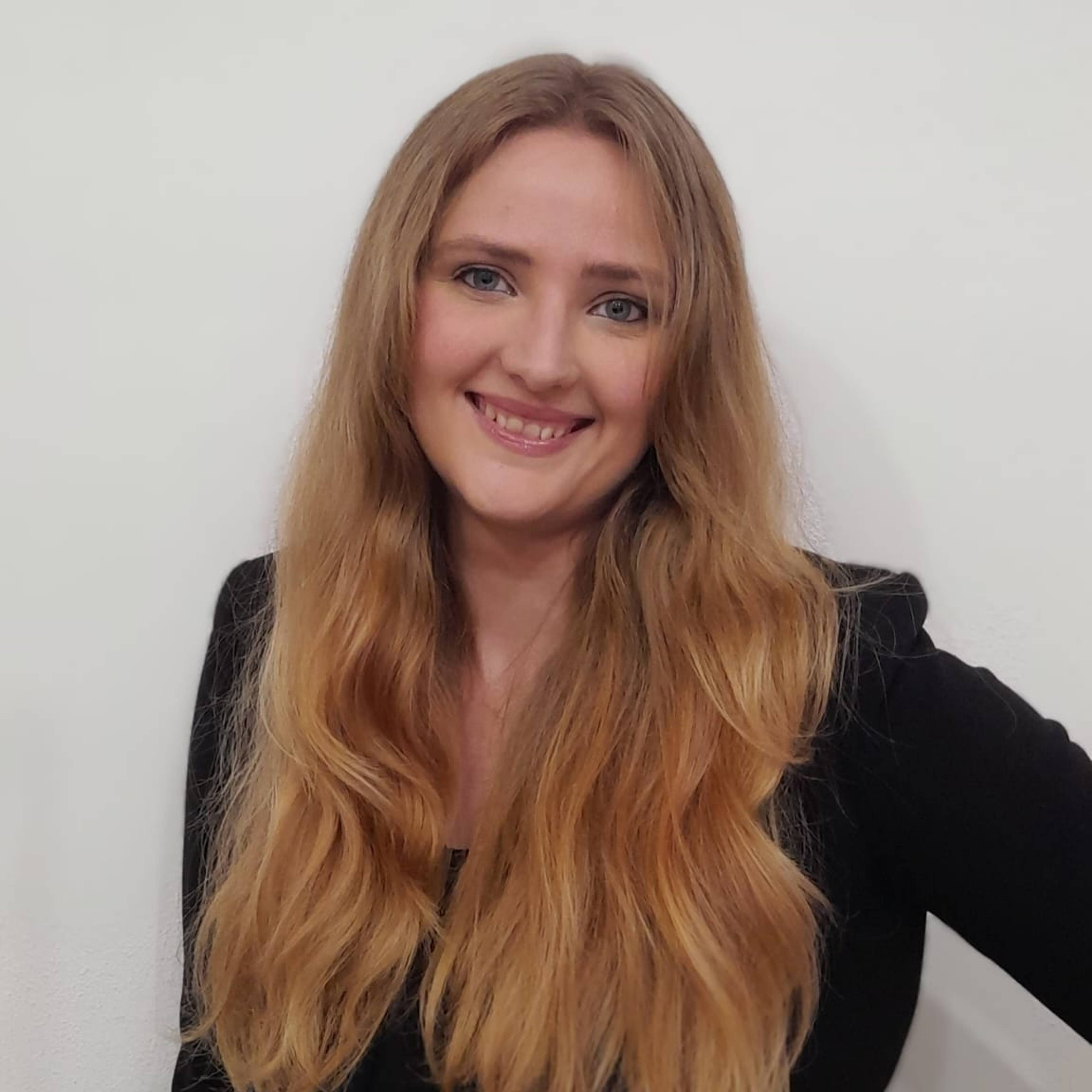 A person with long, wavy blonde hair is smiling warmly at the camera. They are wearing a black top and standing against a plain white background.