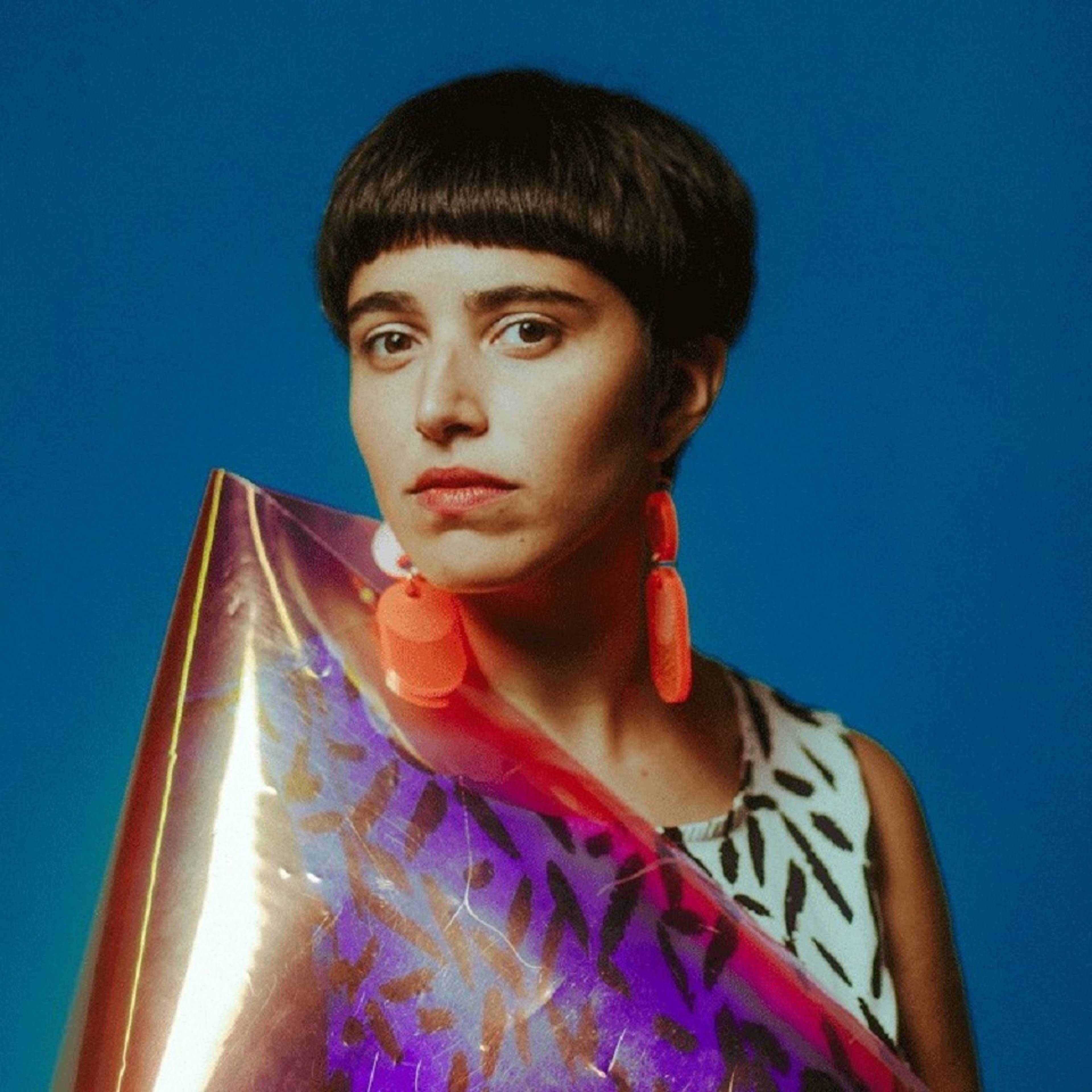 A person with short dark hair and bold earrings poses against a blue background, holding a shiny, colorful reflective material with purple and gold tones. They wear a patterned top.