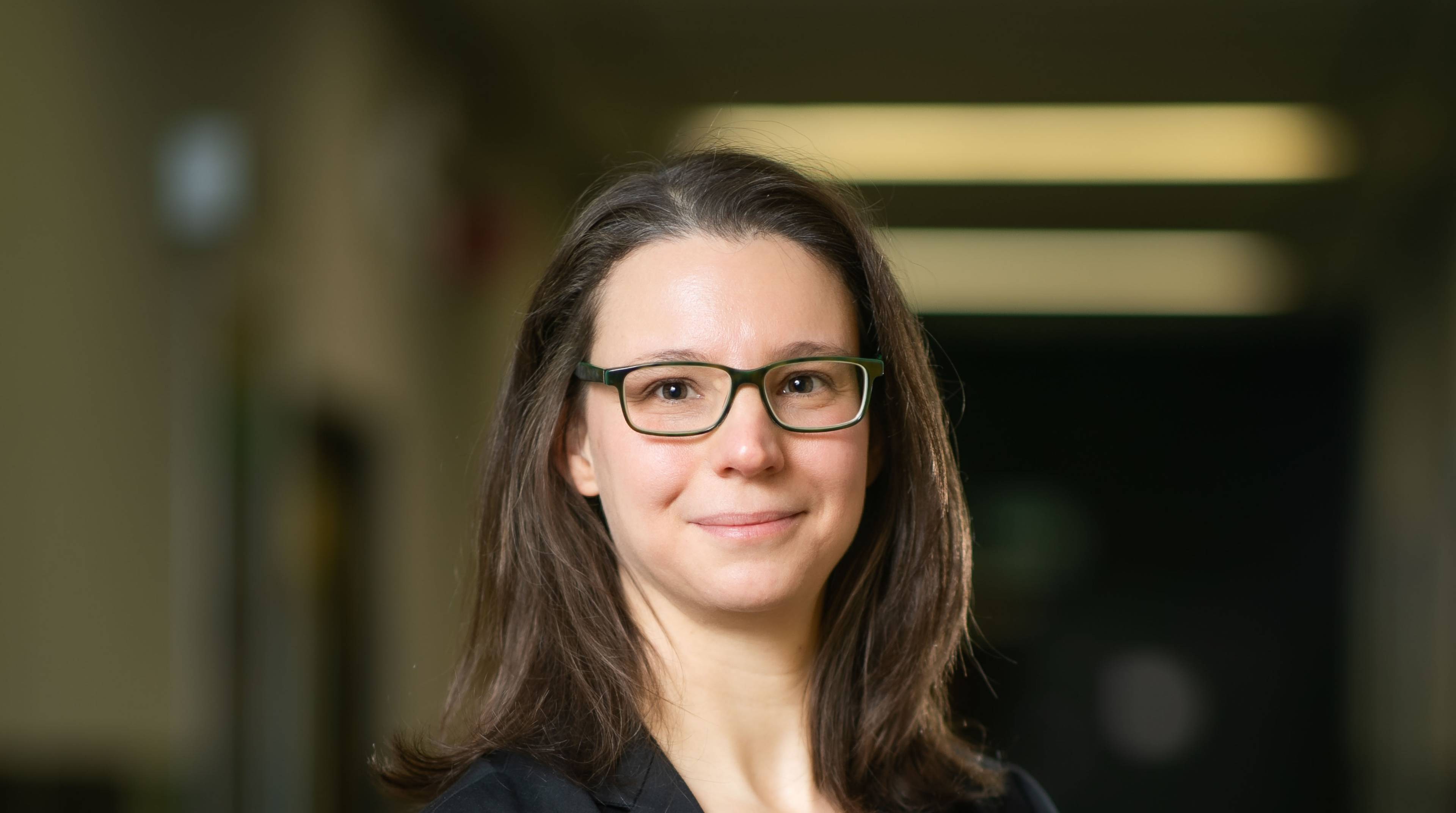 Porträt einer lächelnden Person mit langen braunen Haaren und Brille, aufgenommen in einem Innenraum mit unscharfem Hintergrund.