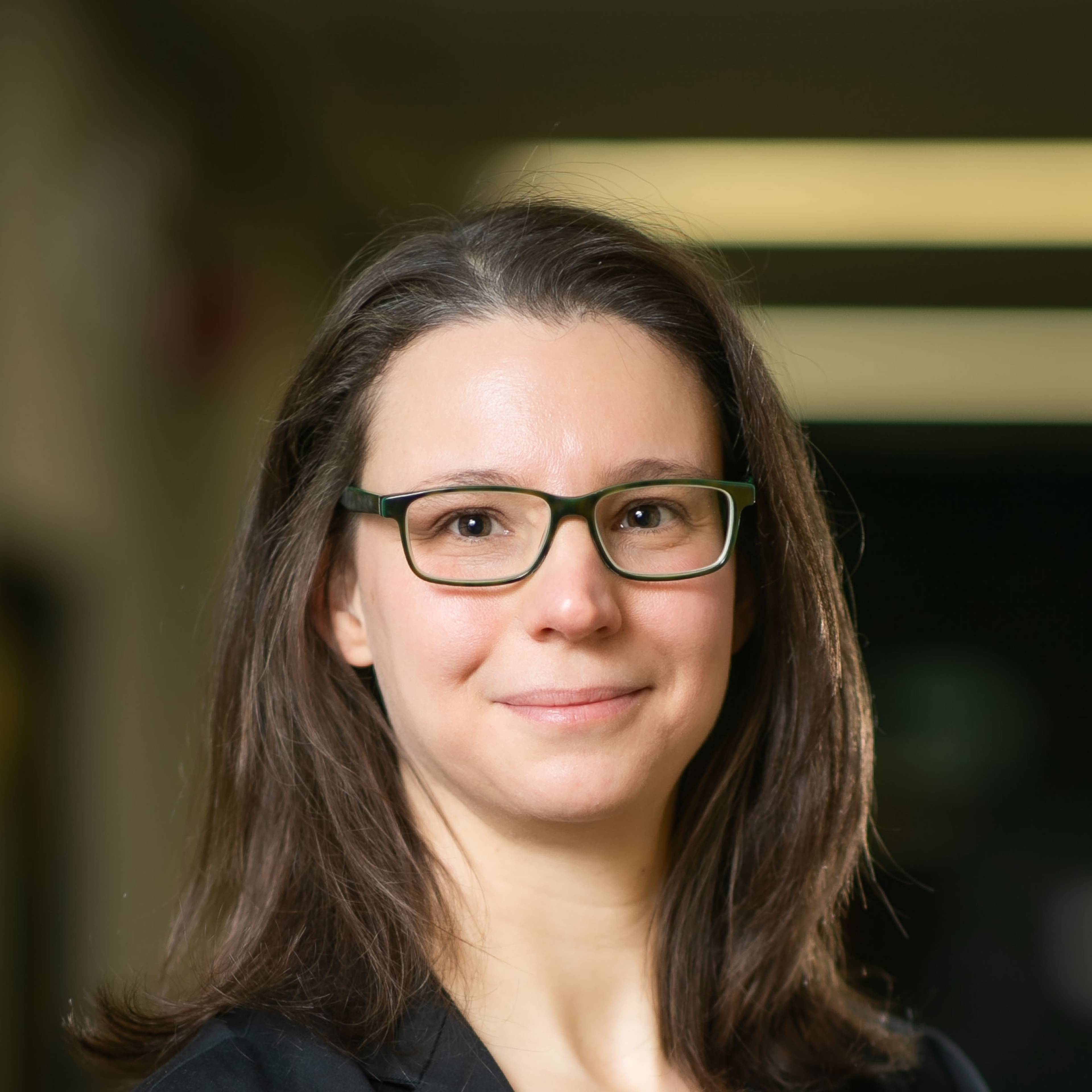 Portrait of a smiling person with long brown hair and glasses, taken indoors with a blurred background.