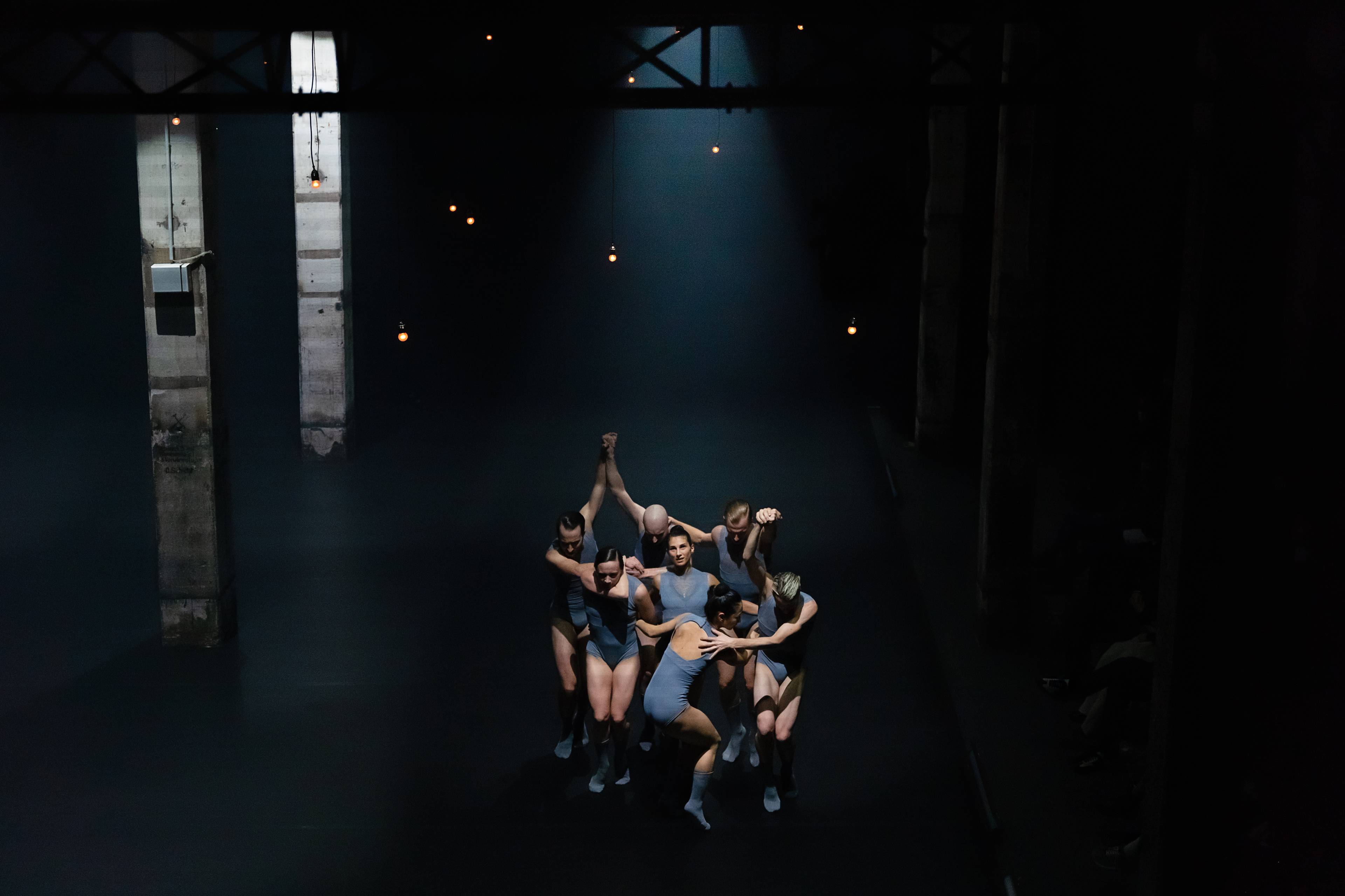 Group of six dancers embracing under a dramatic spotlight in This Is Not a Love Show at Kraftwerk Berlin.