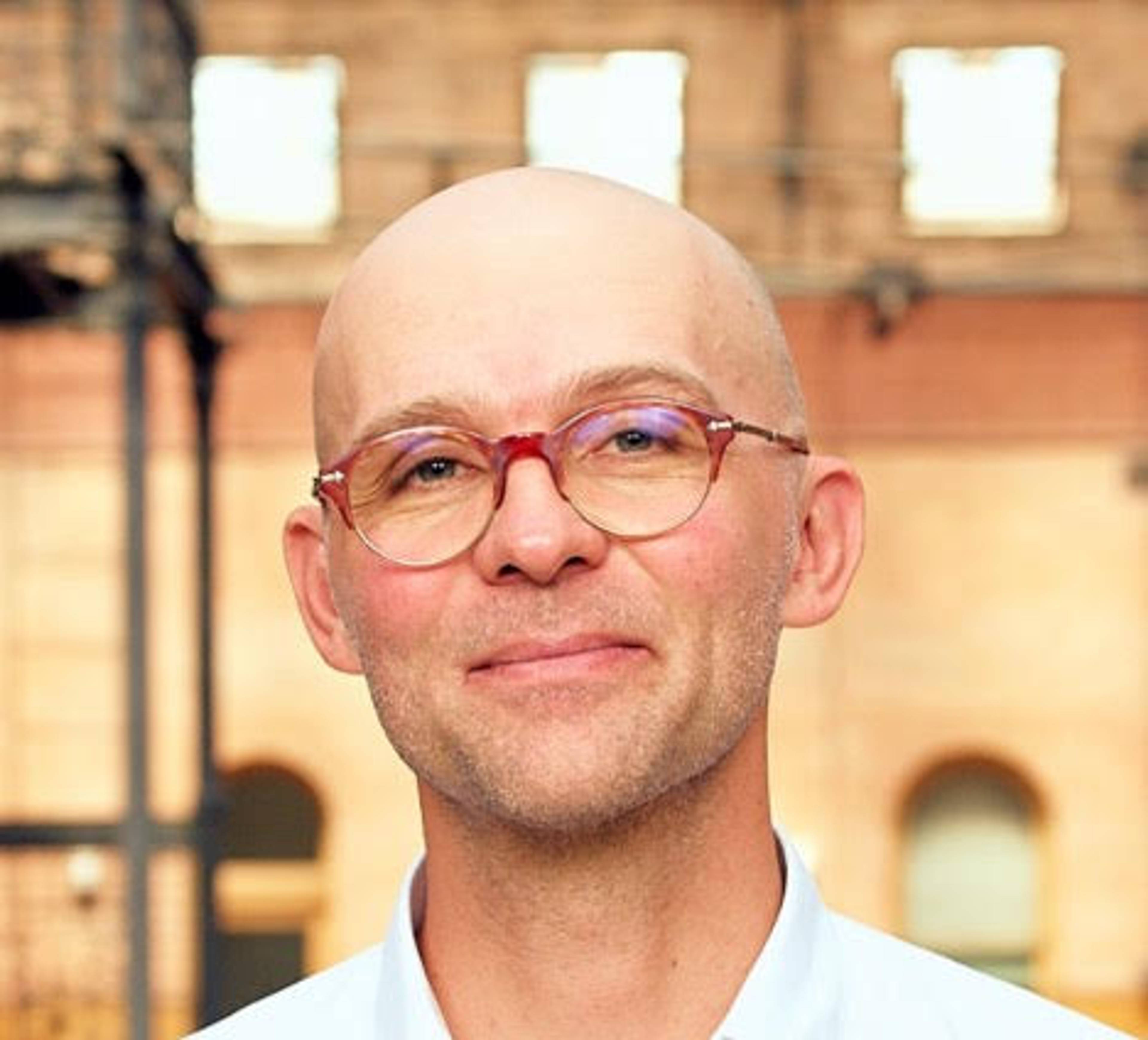 Portrait of a smiling, bald person wearing glasses, standing in front of a historic brick building in daylight.