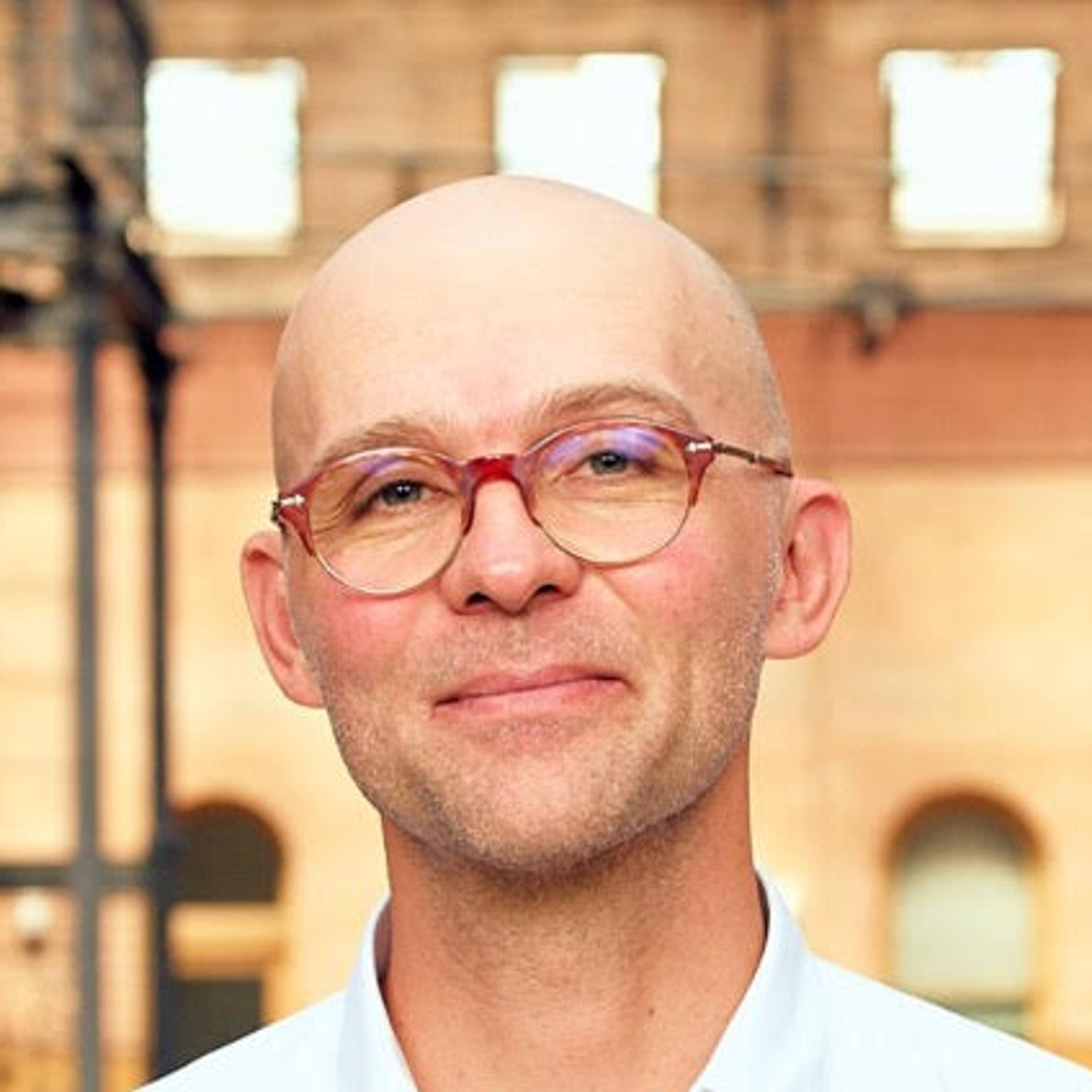 Portrait of a smiling, bald person wearing glasses, standing in front of a historic brick building in daylight.