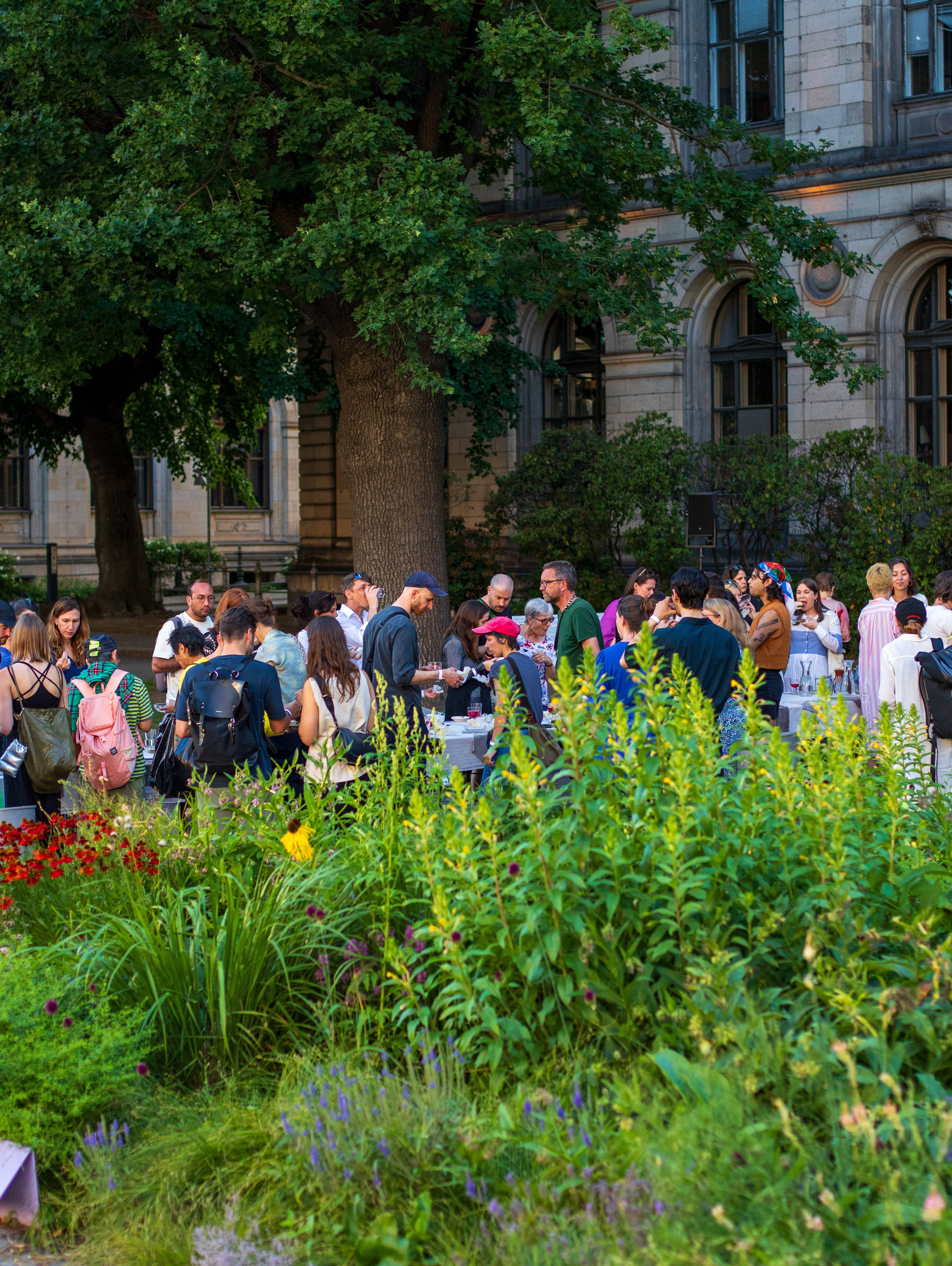 People gathering in front of a historic building beside a lush garden during an outdoor event.