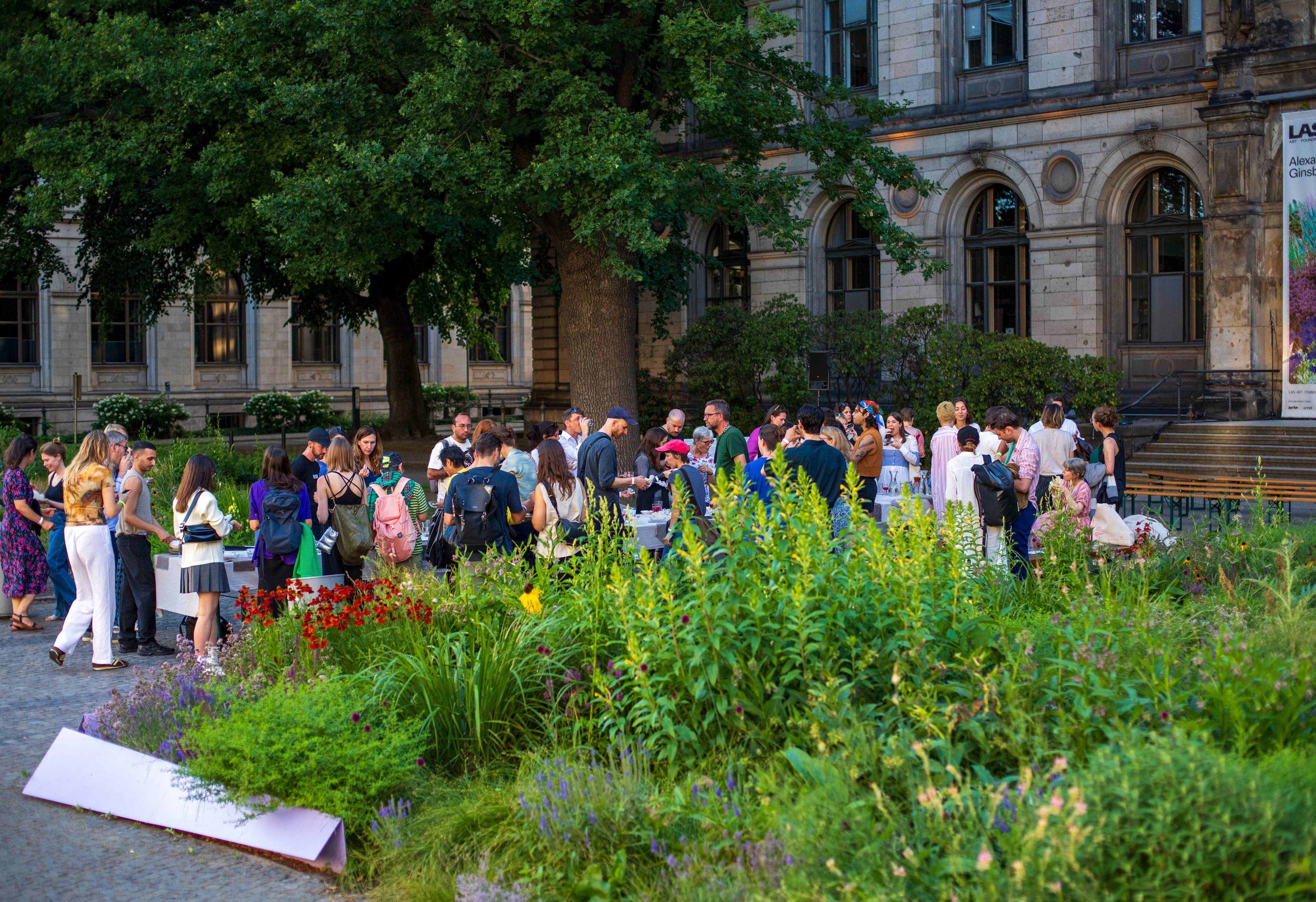 People gathering in front of a historic building beside a lush garden during an outdoor event.