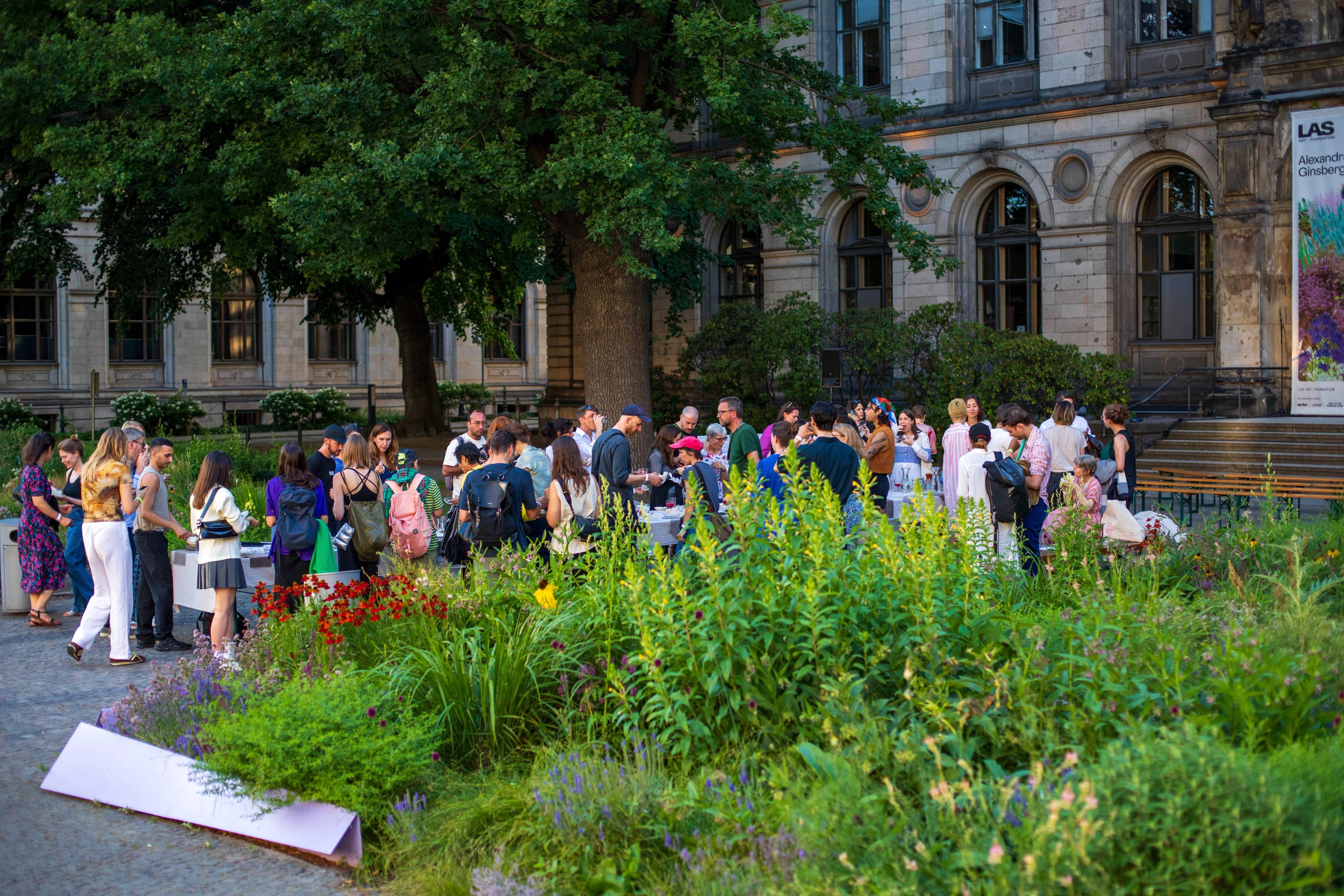People gathering in front of a historic building beside a lush garden during an outdoor event.