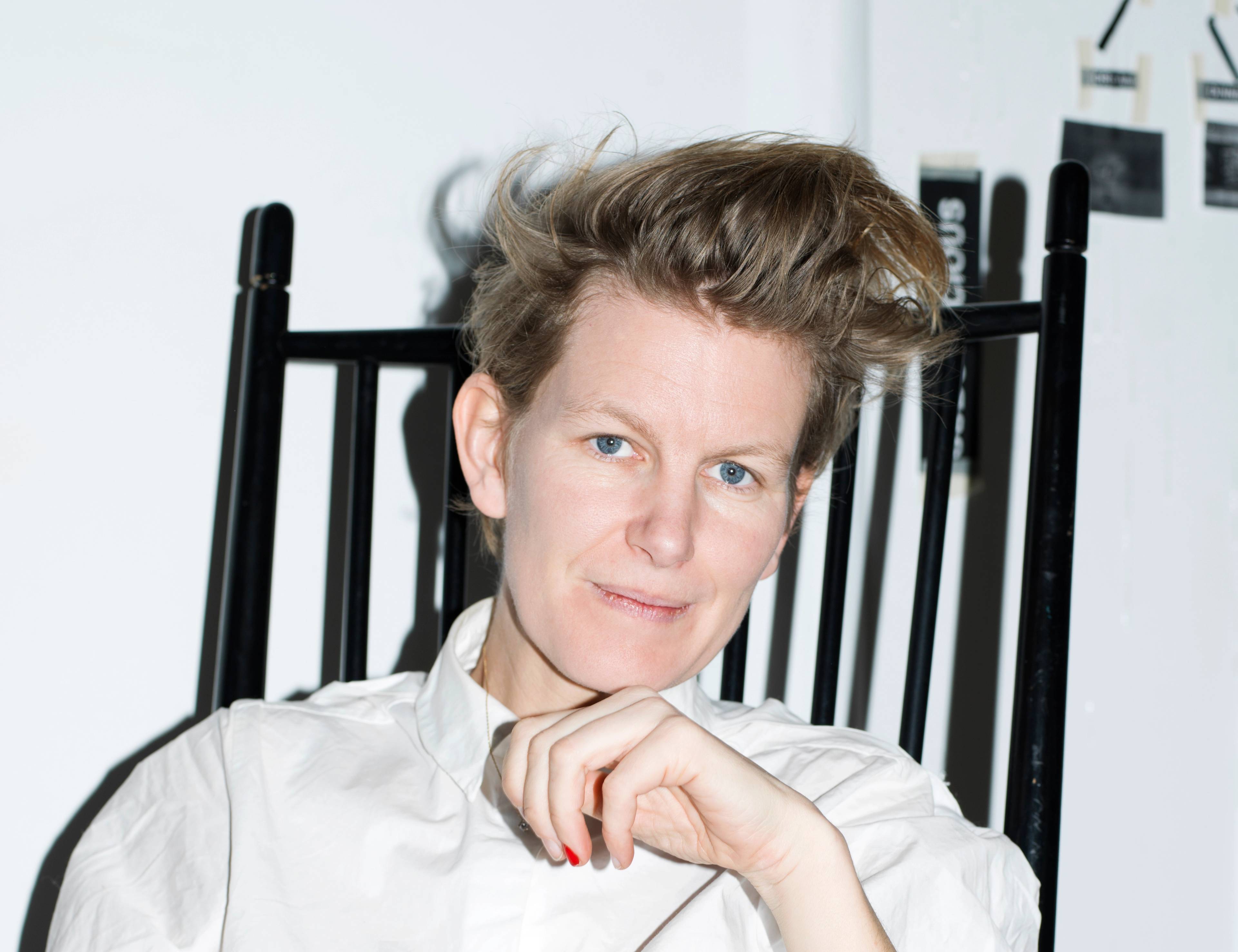 A woman with short, styled hair sits on a black chair, wearing a white shirt. They have a thoughtful expression and rest their hand under their chin.
