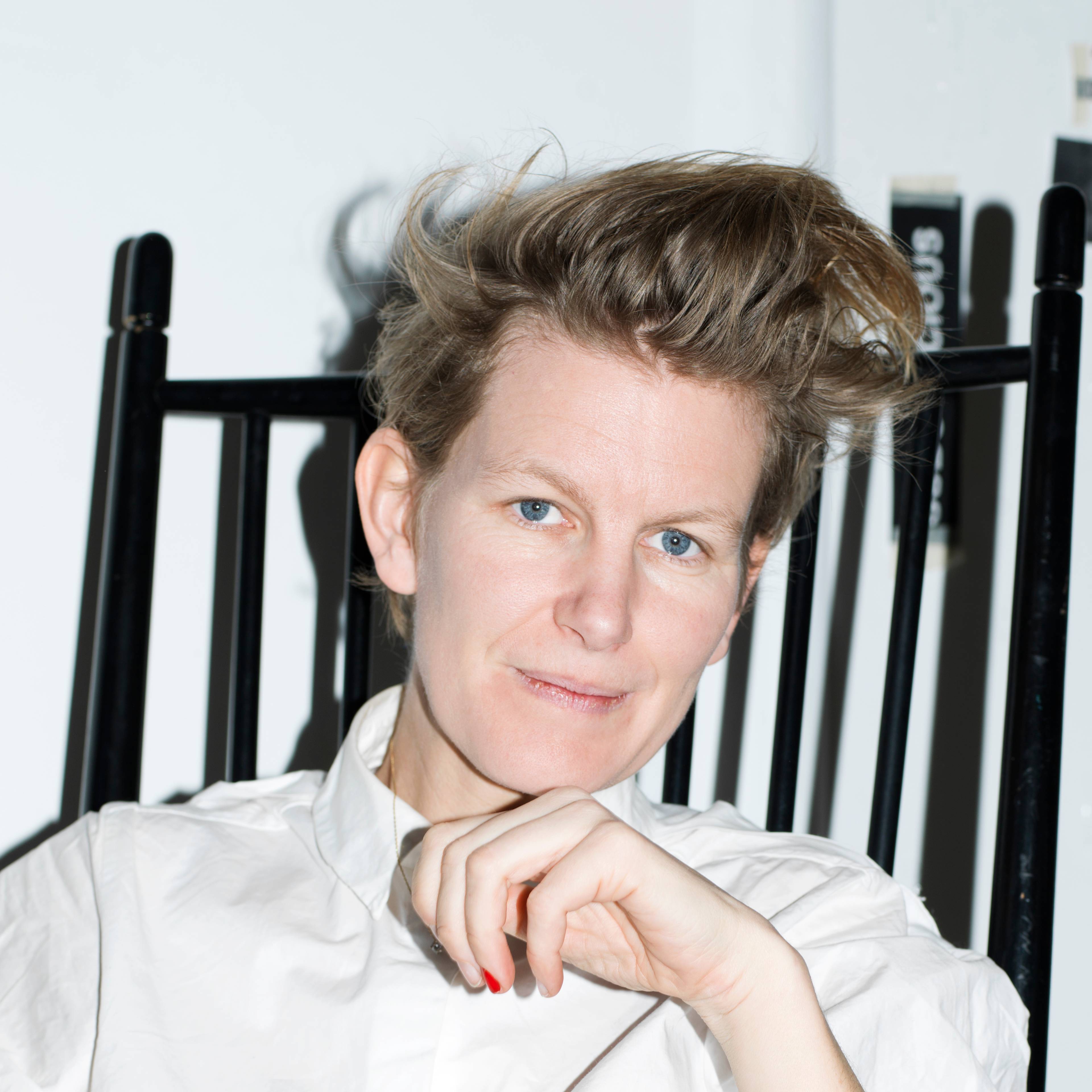 A woman with short, styled hair sits on a black chair, wearing a white shirt. They have a thoughtful expression and rest their hand under their chin.