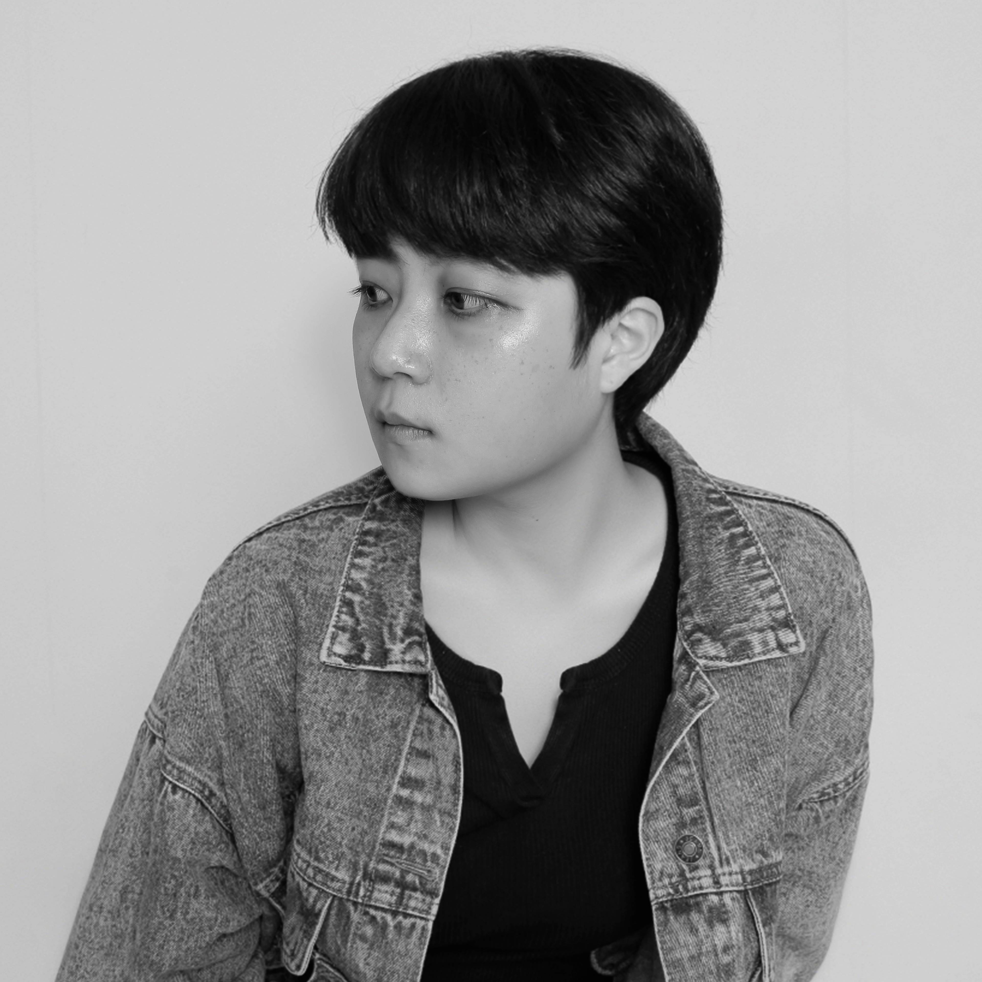A person with short hair looks to the side, wearing a denim jacket over a dark top. The image is in black and white.