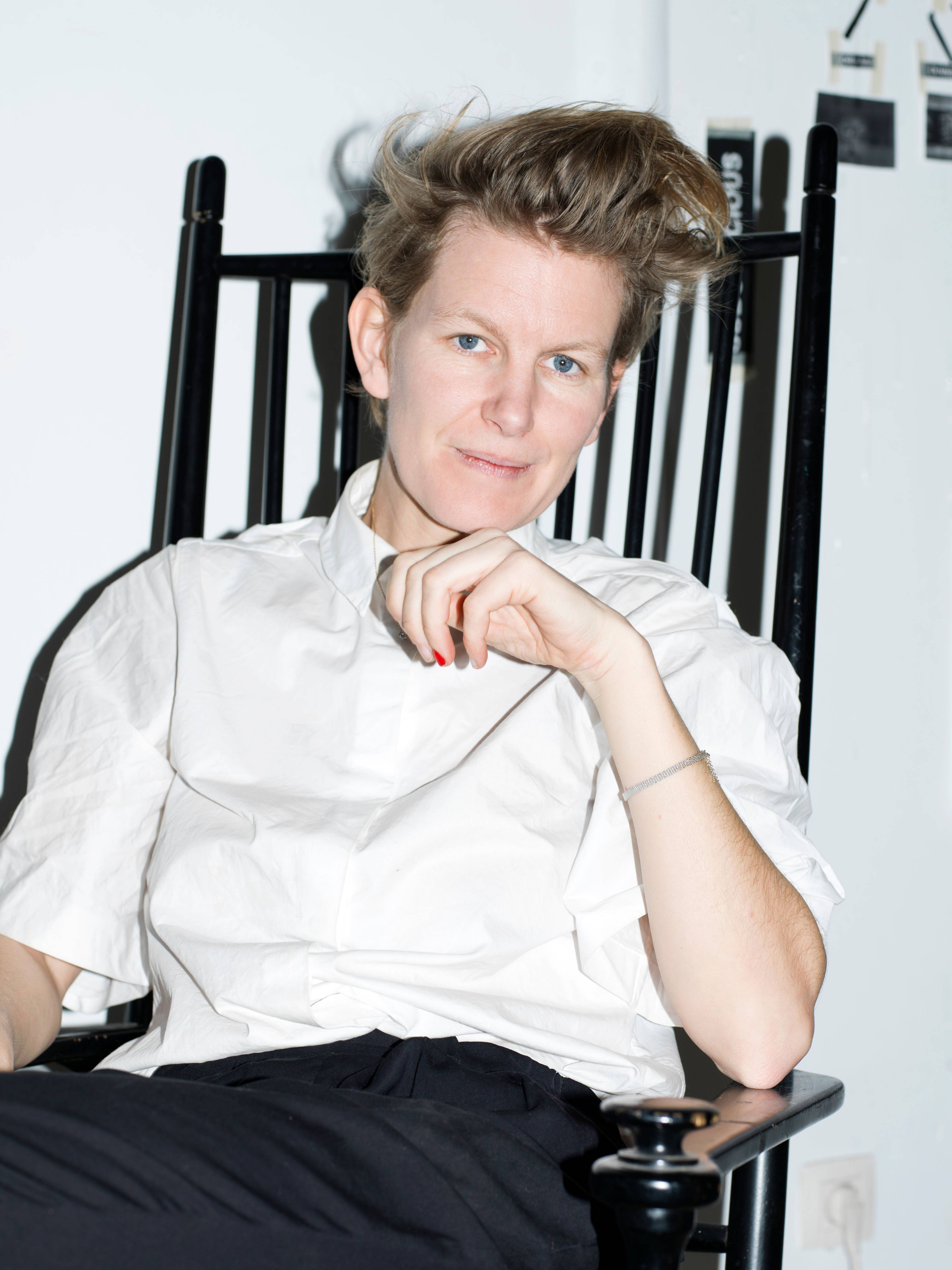 A woman with short, styled hair sits on a black chair, wearing a white shirt. They have a thoughtful expression and rest their hand under their chin.