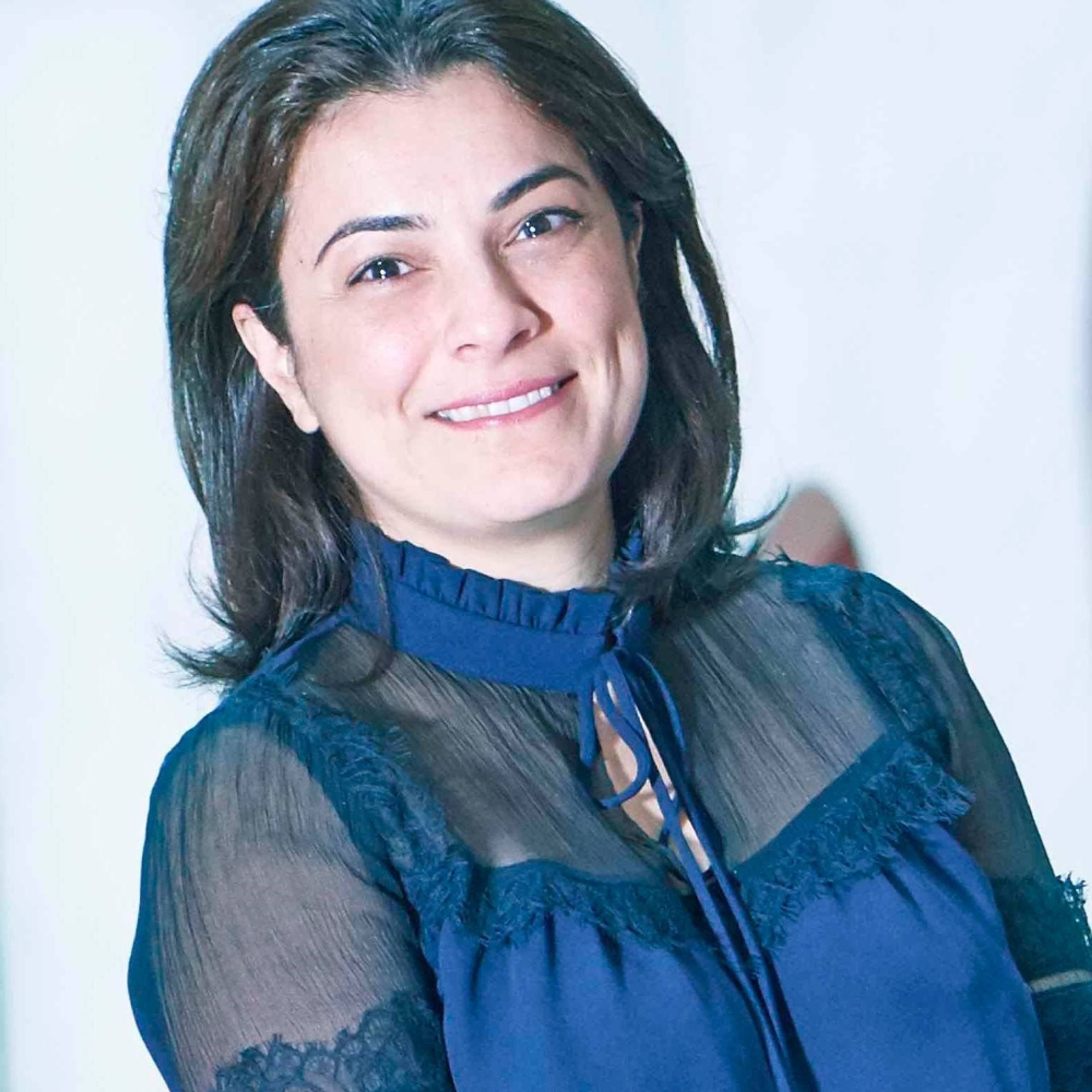 A person with medium-length dark hair smiles at the camera, wearing a dark blue blouse with lace details, standing against a light background.