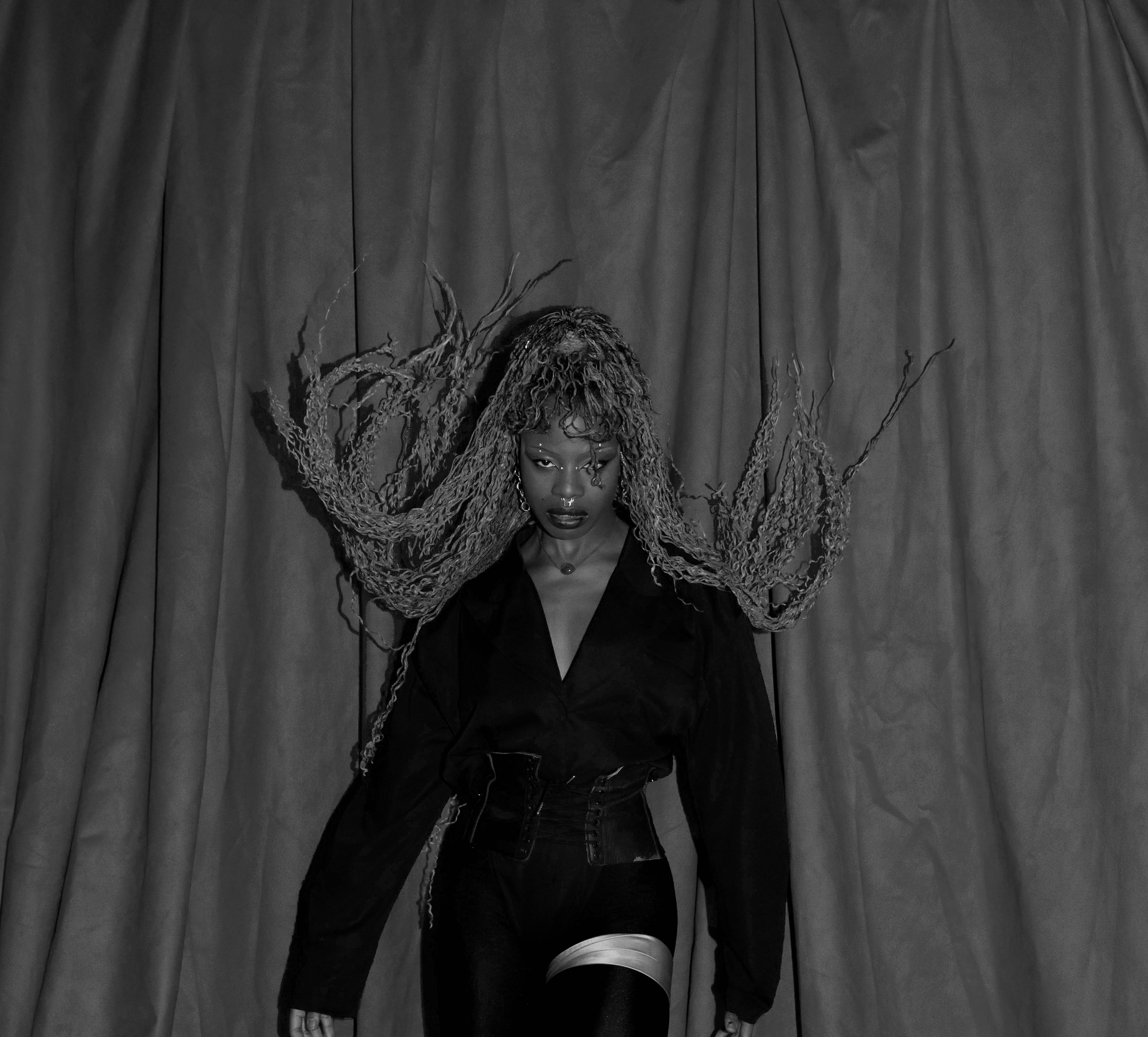 A person stands against a draped curtain, wearing a dark outfit with long, textured hair extending outward. The setting is dramatic with a black and white color scheme, highlighting the pose and expression.