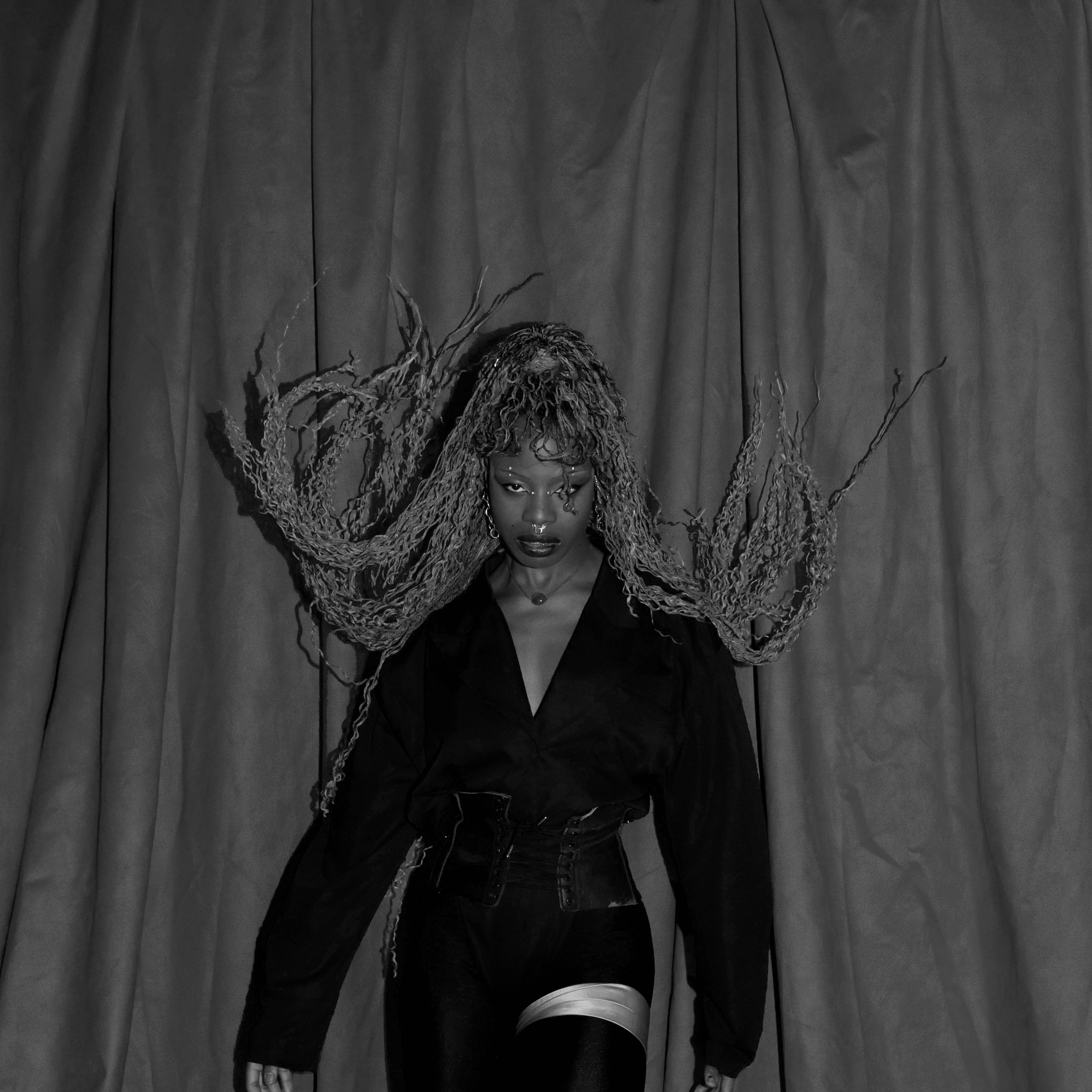 A person stands against a draped curtain, wearing a dark outfit with long, textured hair extending outward. The setting is dramatic with a black and white color scheme, highlighting the pose and expression.