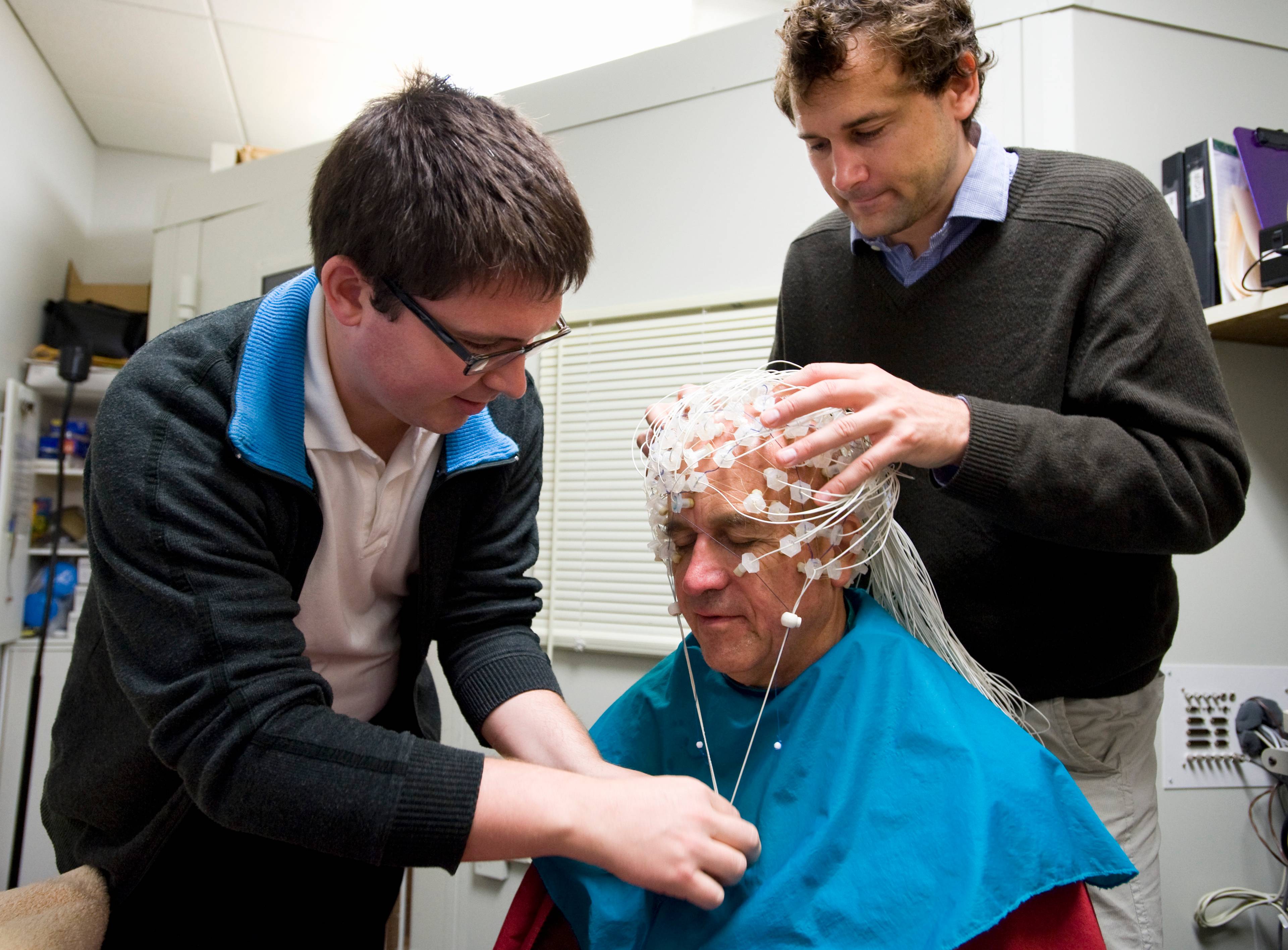 Zwei Männer setzen einem älteren Mann vorsichtig ein EEG-Stirnband auf den Kopf.