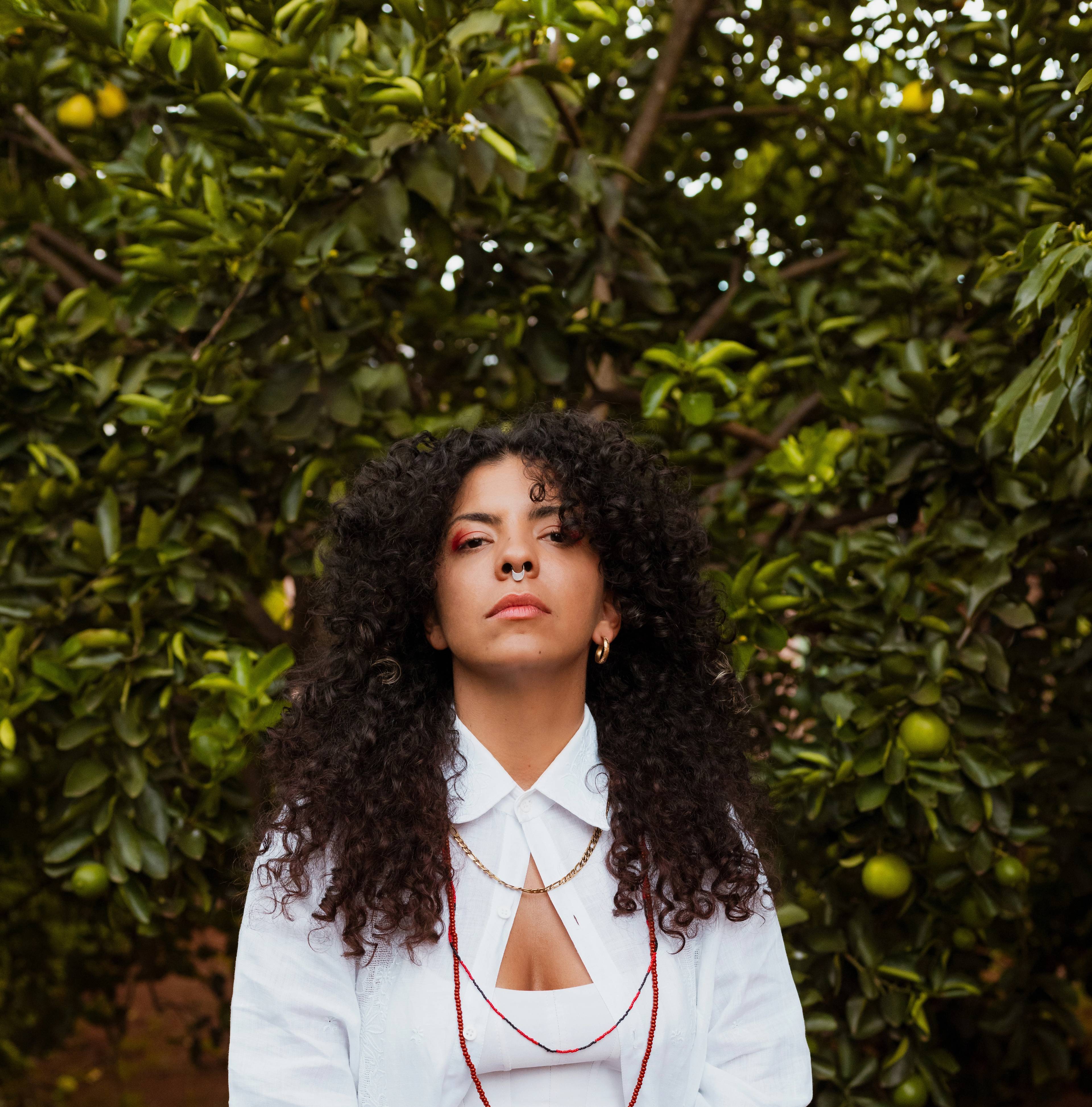 A person with curly hair, wearing a white shirt and red jewelry, stands in front of a lush green orange tree. The person gazes confidently at the camera.
