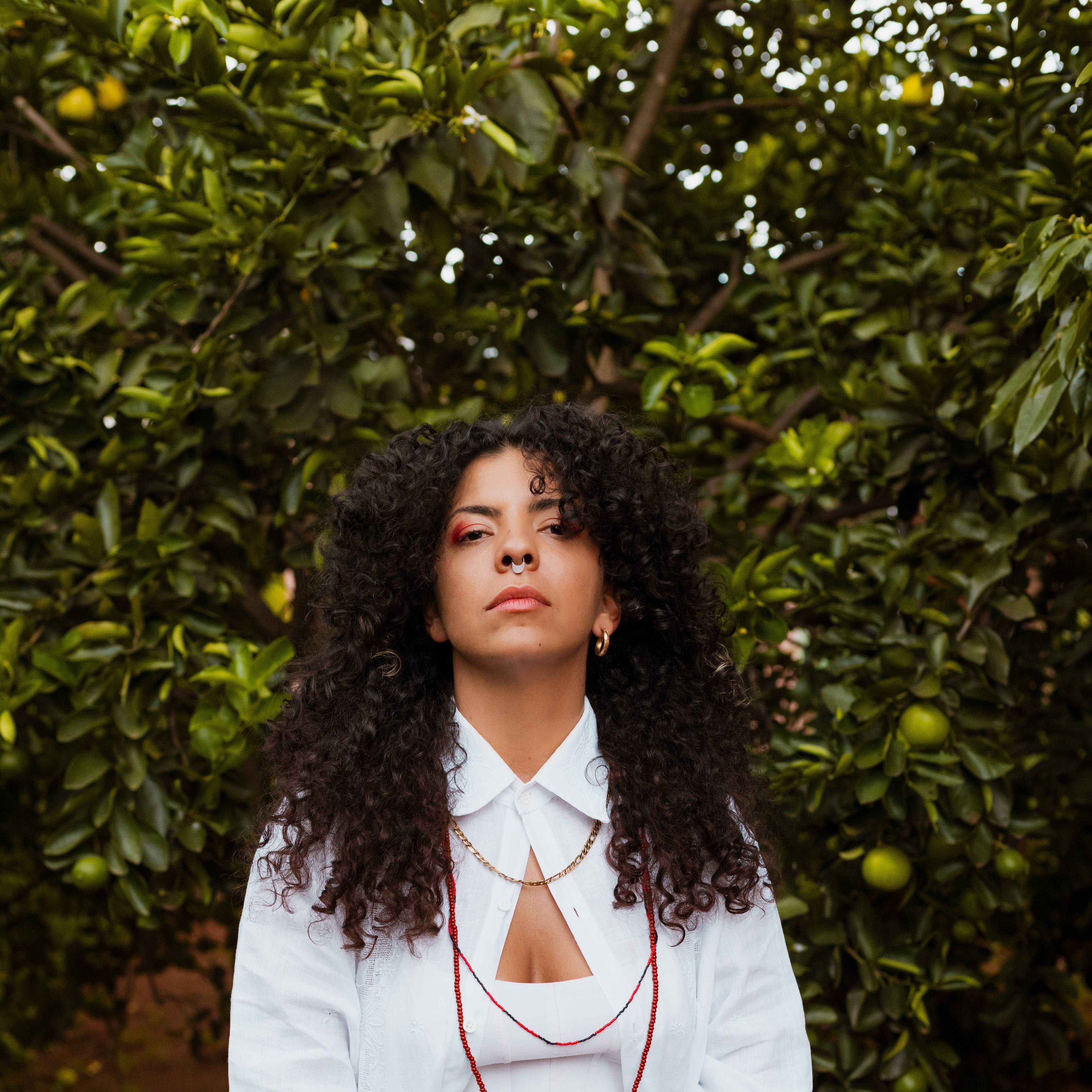 A person with curly hair, wearing a white shirt and red jewelry, stands in front of a lush green orange tree. The person gazes confidently at the camera.